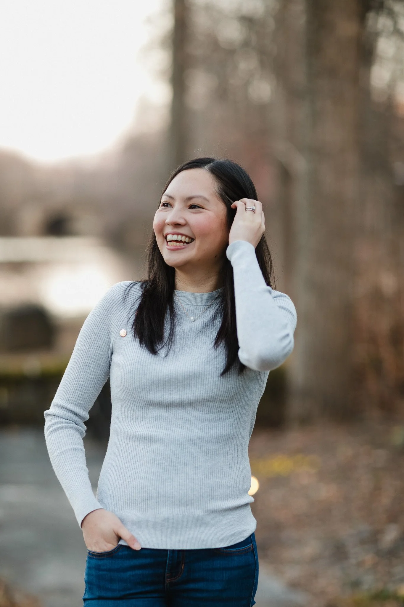 A young woman with long dark hair smiling and wearing a light gray sweater, standing outdoors in a park or wooded area in autumn.