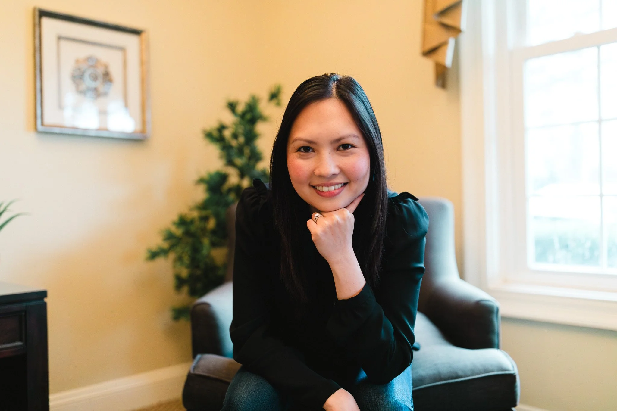 A woman with long black hair sitting in a black leather chair, smiling with her chin resting on her hand, in a warmly lit room with a window and a framed picture on the wall behind her. Bing Gonzalez Coaching. Professional Neuroscience Coach.