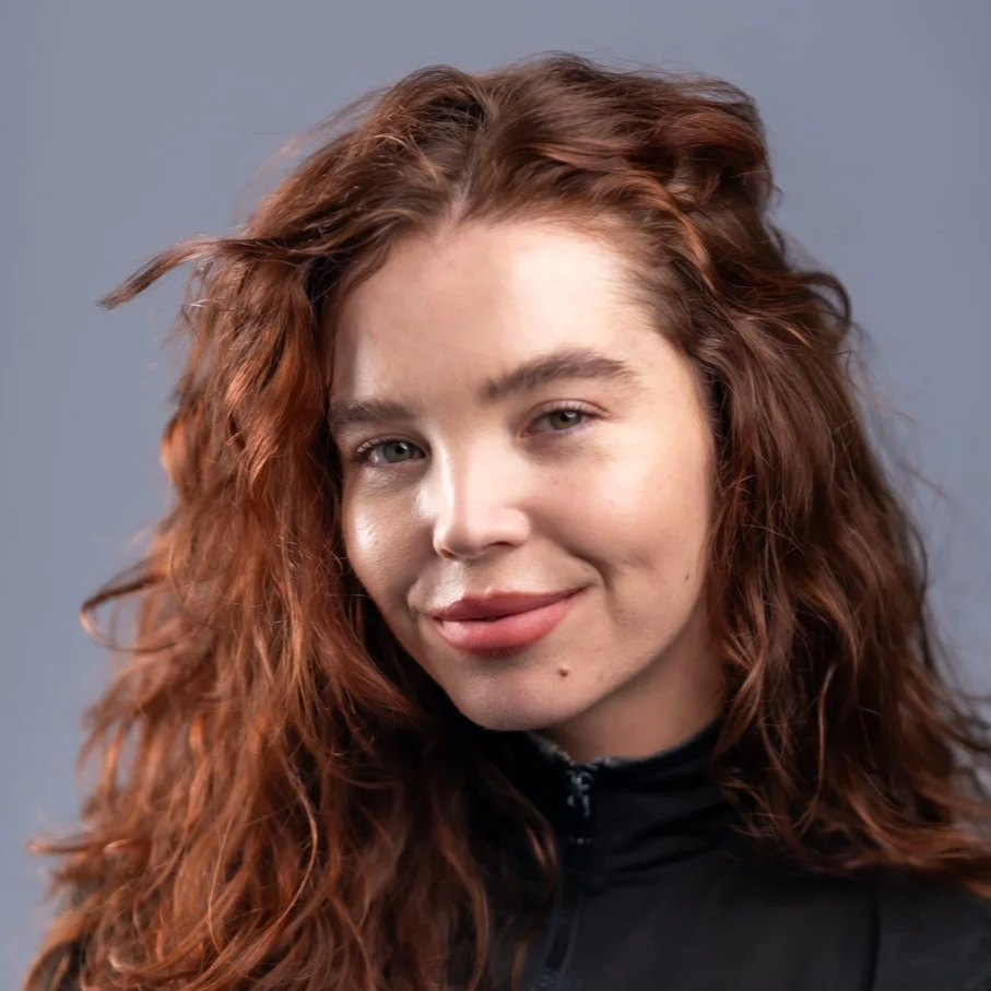 Close-up portrait of a young woman with wavy red hair, light skin, and a subtle smile, wearing a black zip-up jacket against a gray background.