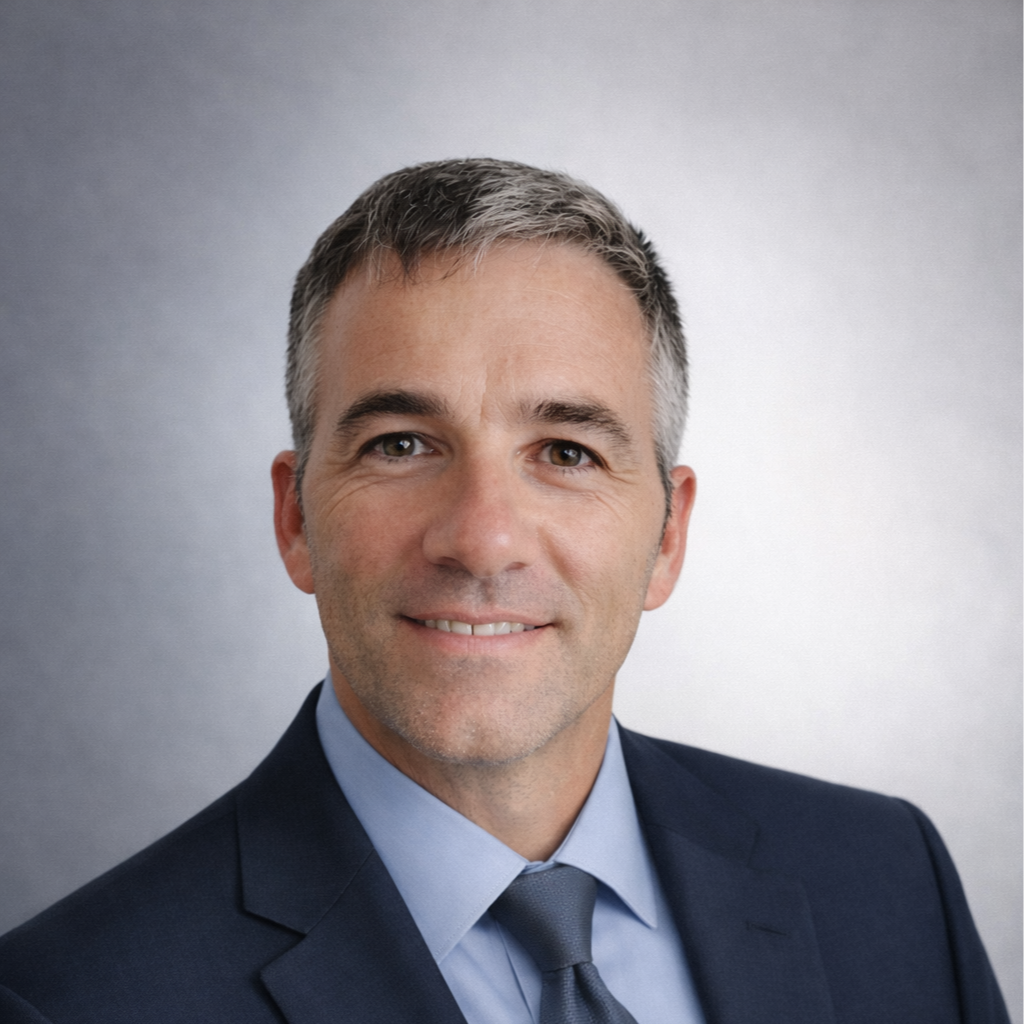 A professional headshot of a middle-aged man with short gray hair, wearing a dark suit, light blue shirt, and a dark tie, smiling slightly against a light gray background.