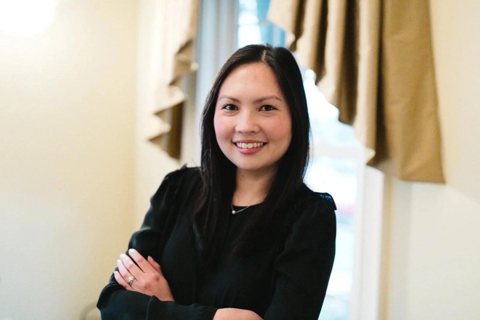 A woman with long black hair, smiling, wearing a black top, standing with her arms crossed, in front of a window with gold-colored curtains. Bing Gonzalez. Professional Neuroscience Coach.