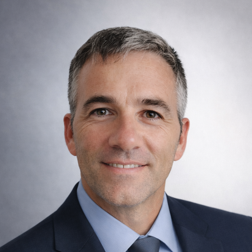 Headshot of a middle-aged man with short, salt-and-pepper hair, wearing a dark suit, light blue shirt, and dark tie, smiling softly against a gray background.