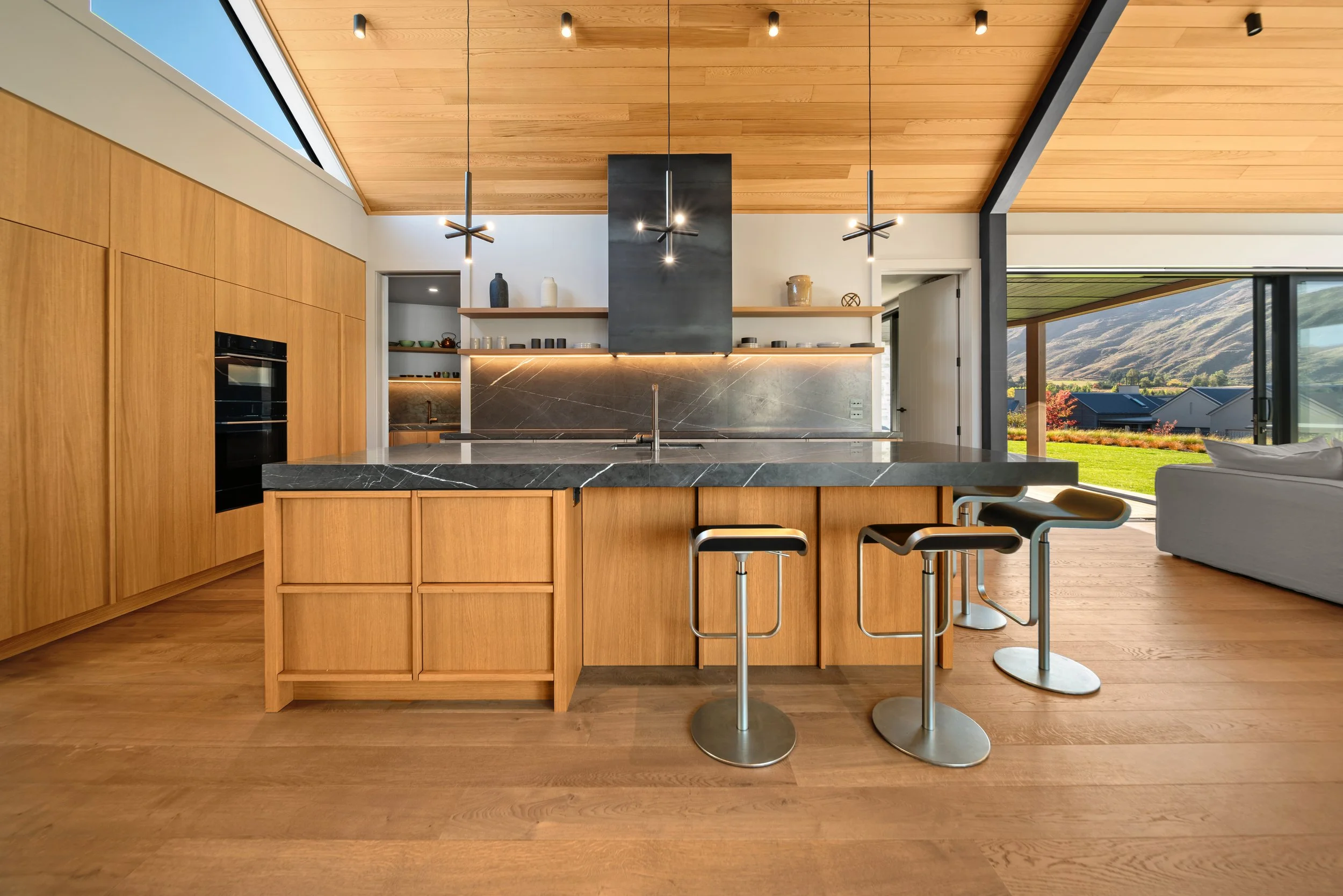 Modern kitchen with wooden cabinets and ceiling, black marble island, three barstools, open to living room with large window showing mountain view.
