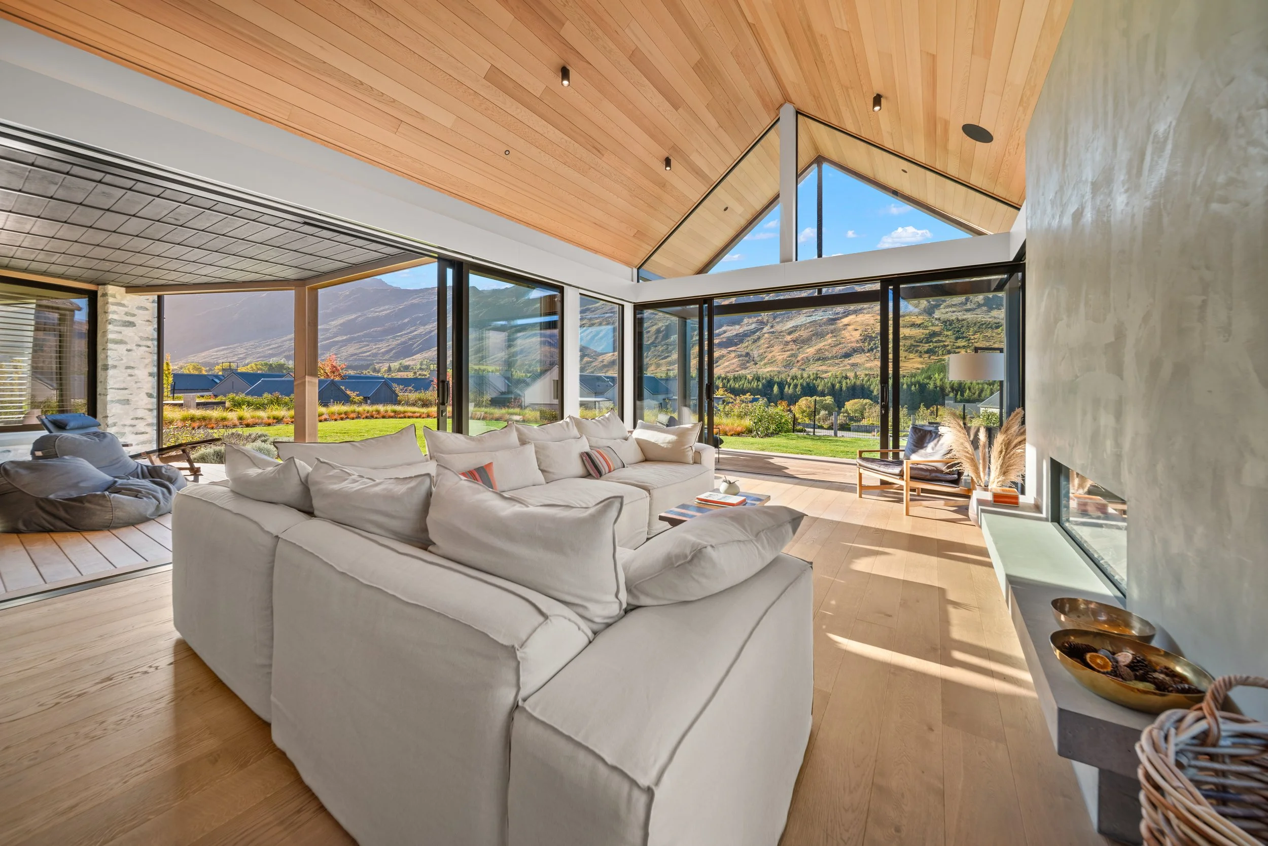 Interior view of a modern living room with large glass windows showing mountains and blue skies outside. White sectional sofa with cushions, wooden flooring, and a ceiling with wooden panels. Various decorative items including a table with books, a lamp, and bowls.