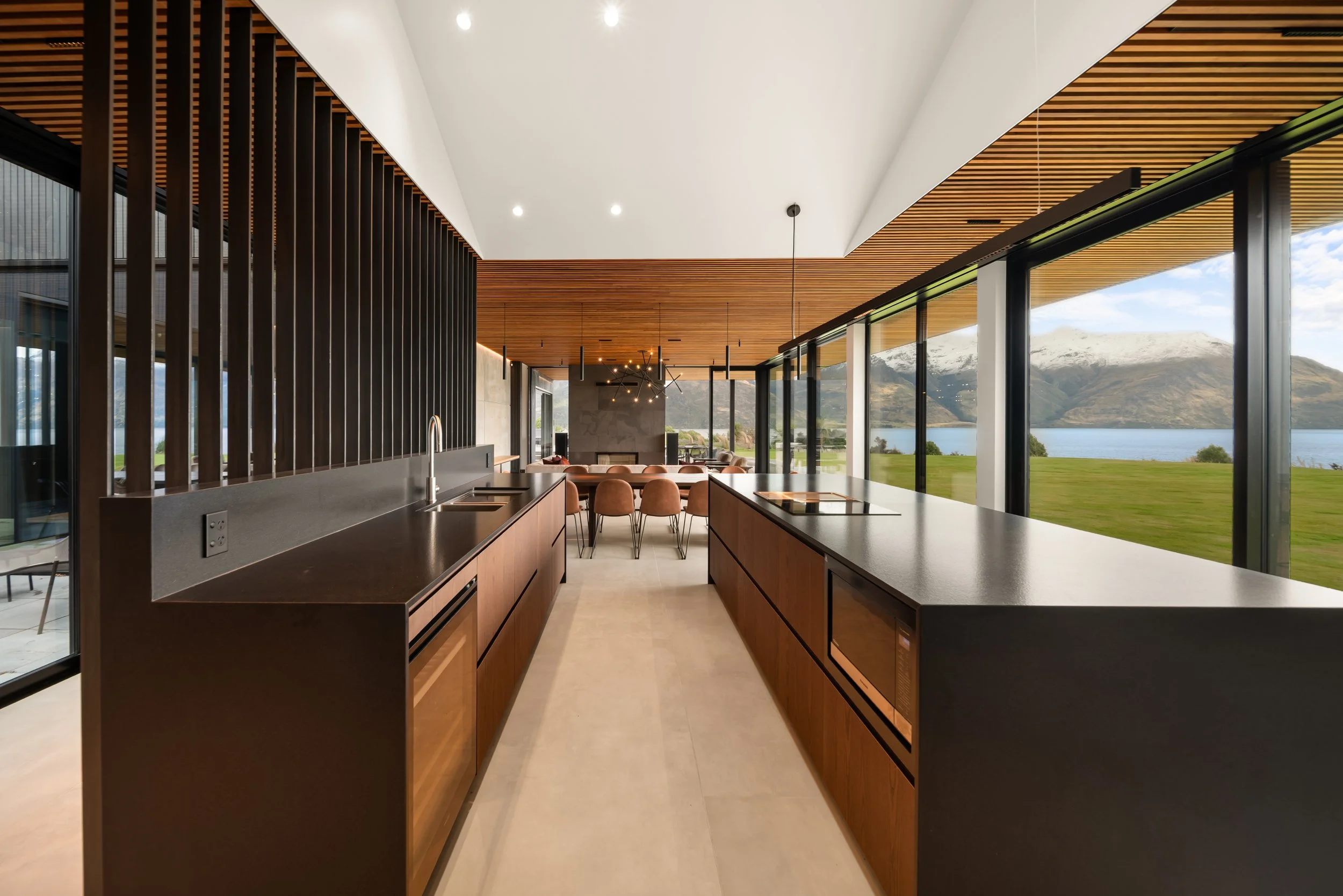Modern kitchen with black countertops, wooden cabinets, and large windows showing mountains and a lake outside.
