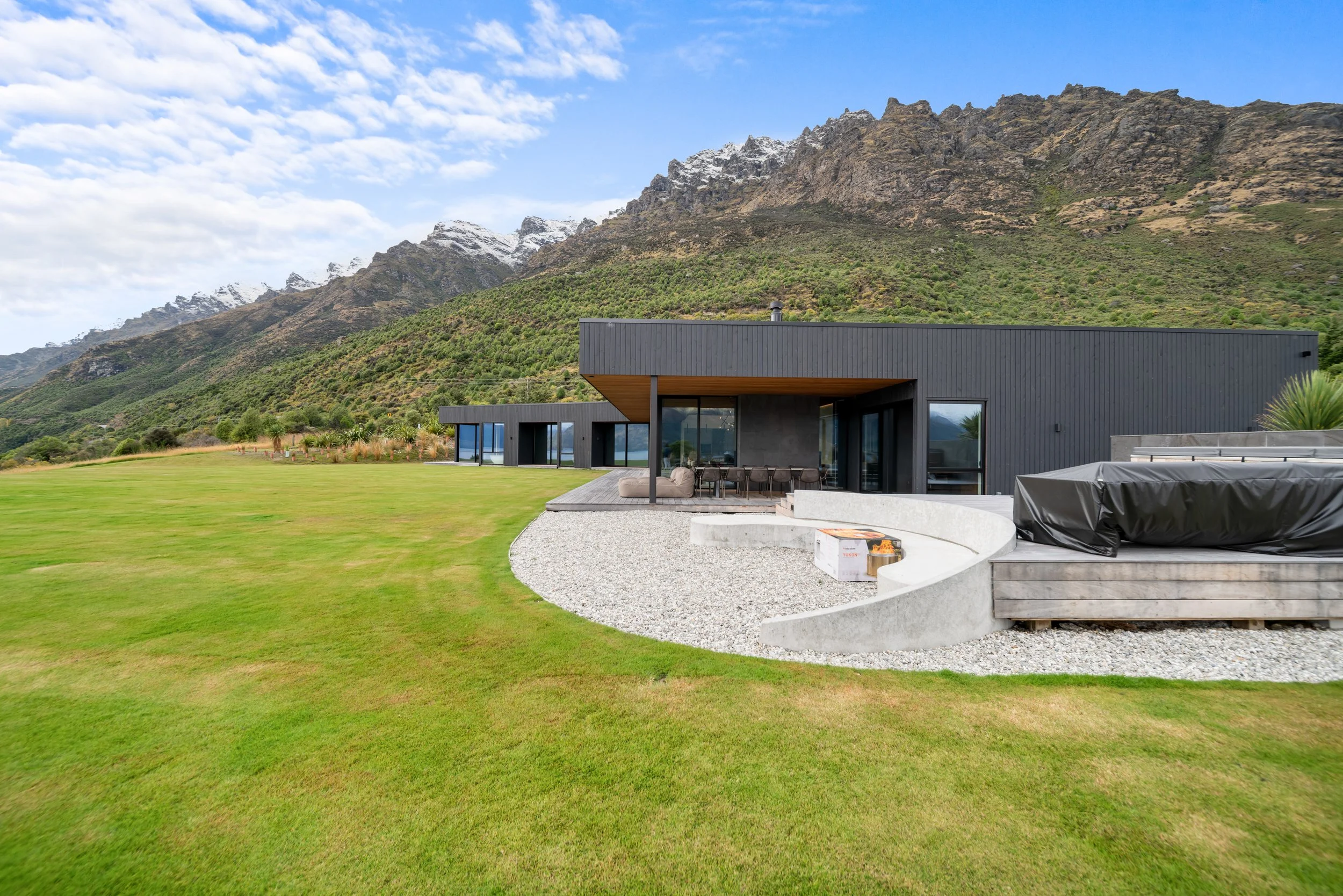 Modern house with large glass windows and dark exterior, situated on a spacious grassy lawn, with a mountain range in the background and a partly cloudy blue sky overhead.