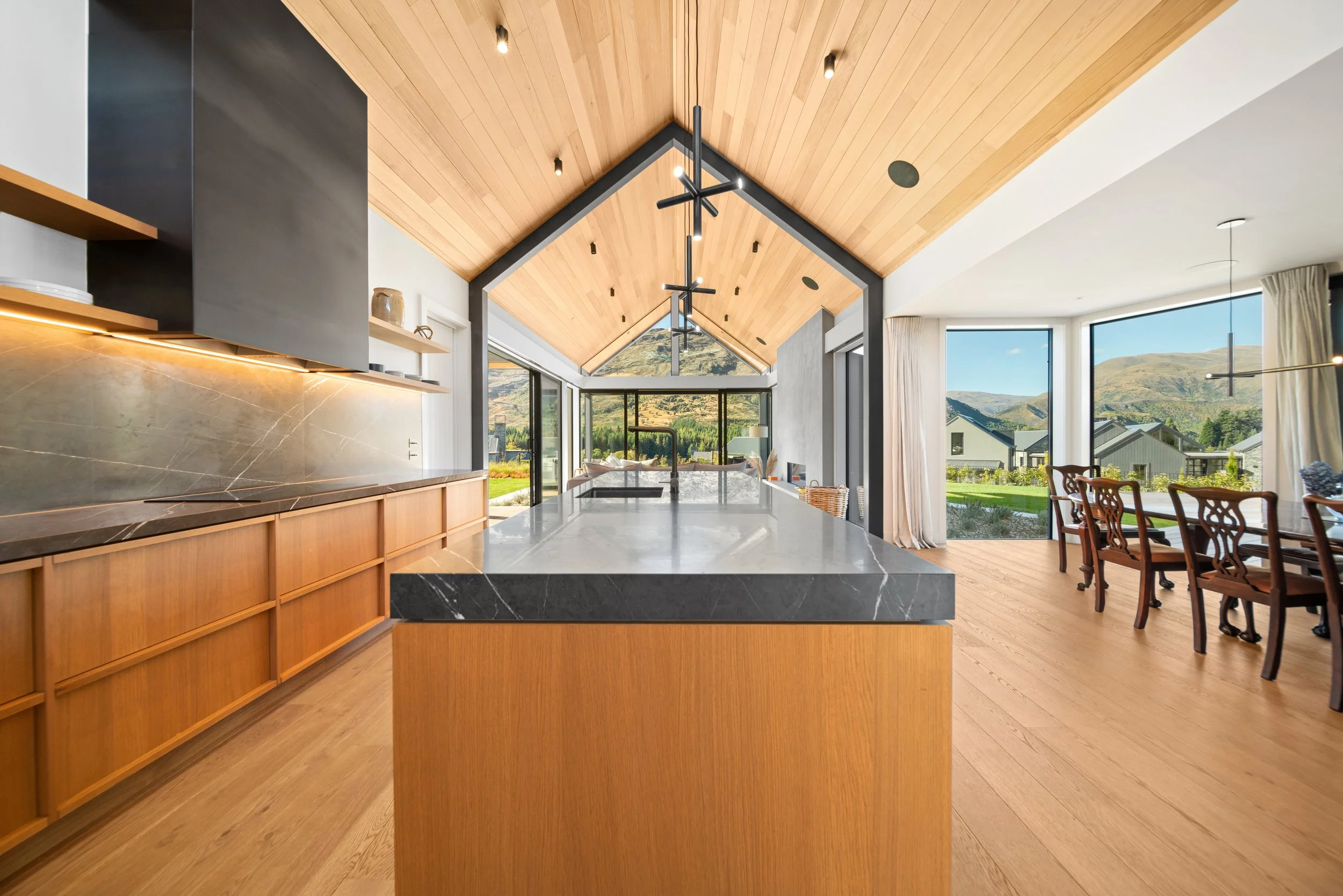 Modern kitchen with wood cabinets, marble island counter, and large windows showing a mountain landscape.