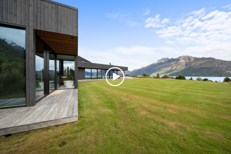 Modern black house with large glass windows overlooking green lawn, mountains, and a body of water, under a partly cloudy sky.