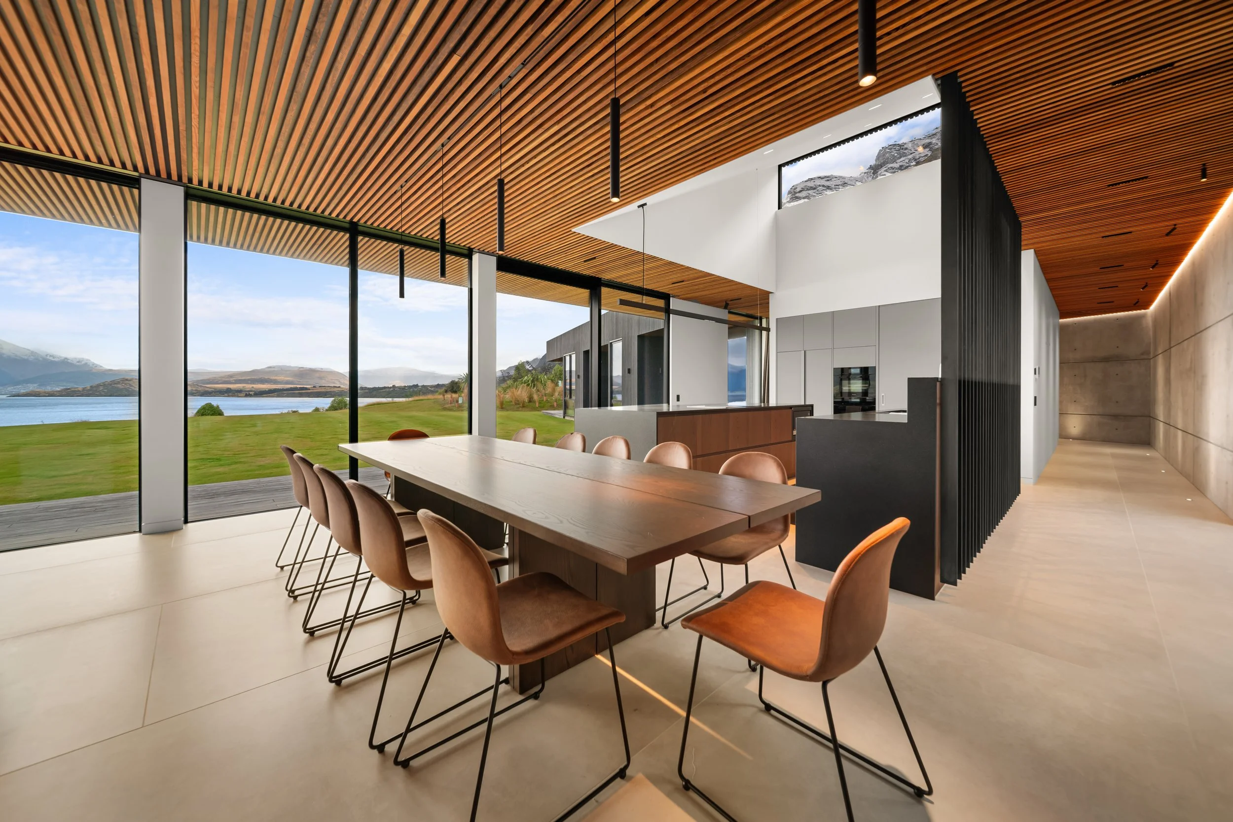 Modern dining room with large windows looking out to a scenic lake and mountains, featuring a long wooden table surrounded by tan chairs, wooden paneled ceiling with hanging black light fixtures, and a combination of white and concrete walls.
