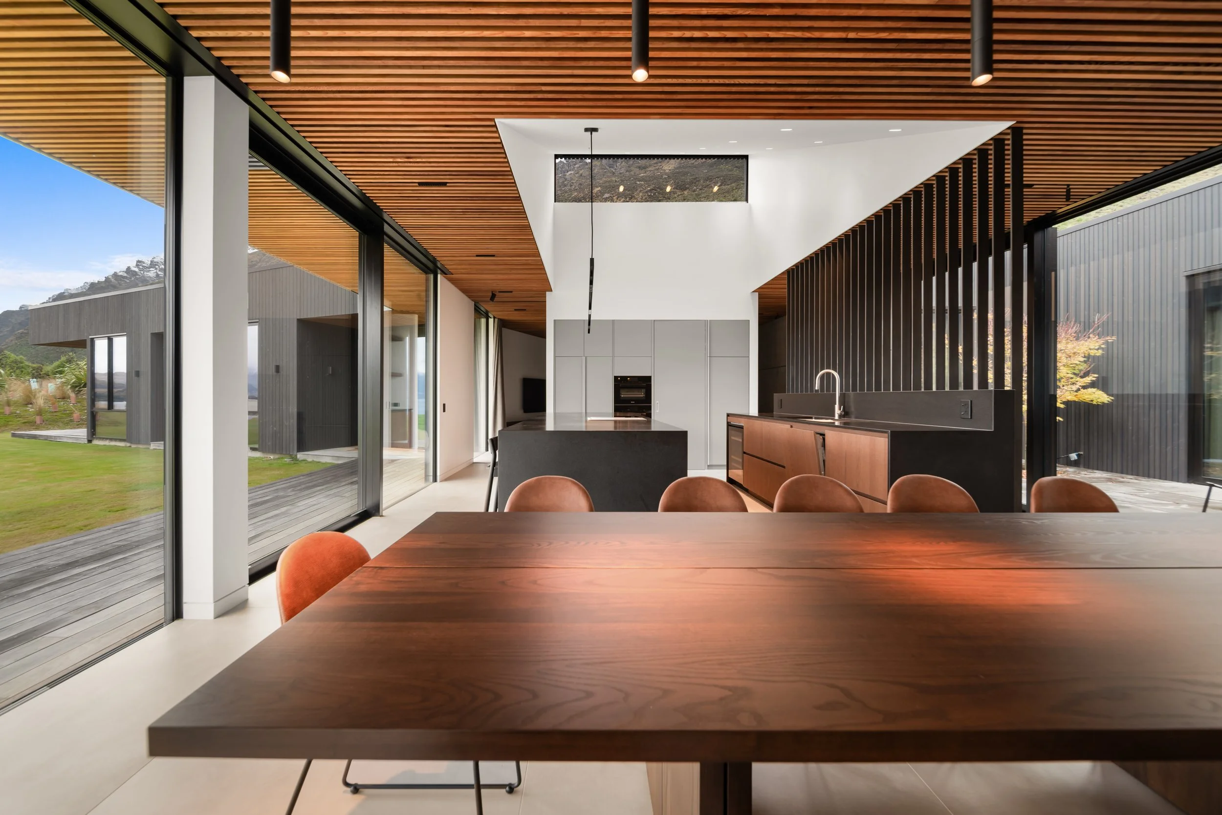 Interior of a modern open-concept kitchen and dining area with a wooden ceiling, large sliding glass doors, and a view of an outdoor yard with mountains in the background. The space features a dark island, brown chairs, and minimalist cabinetry.