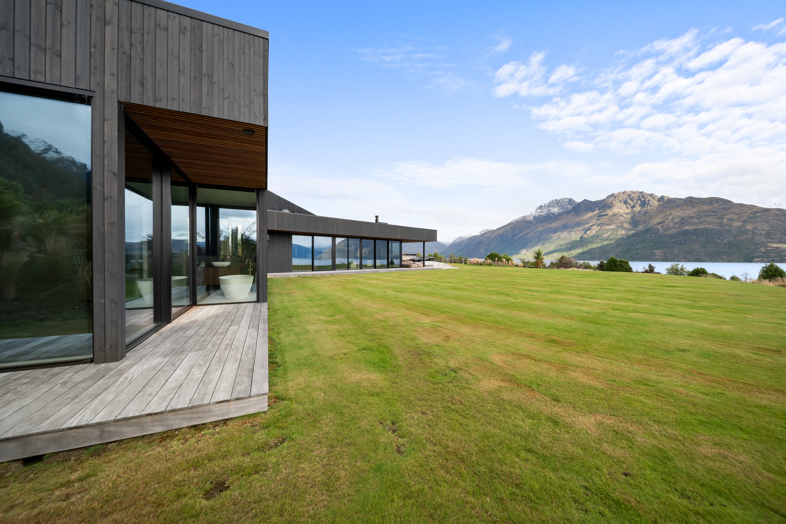 Modern house with large glass windows and wooden exterior, set on a grassy landscape with a mountain and lake in the background under a blue sky with clouds.