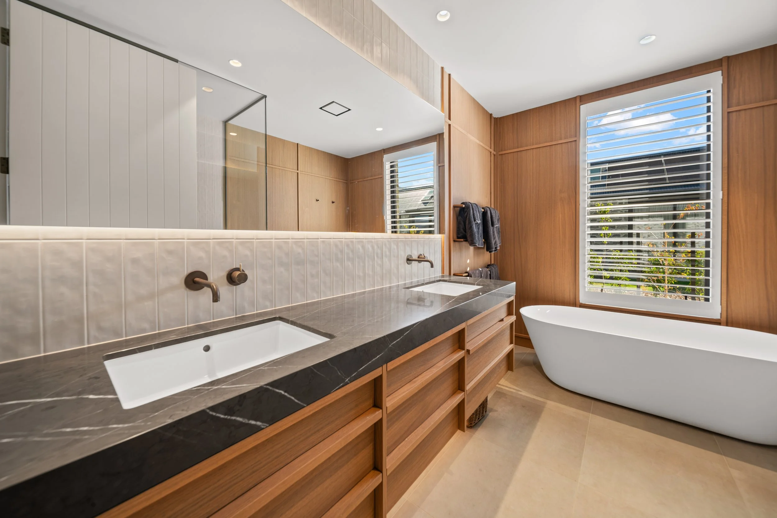 Modern bathroom with a black marble countertop, white vessel bathtub, large window with white blinds, wood-paneled walls, and a mirror.