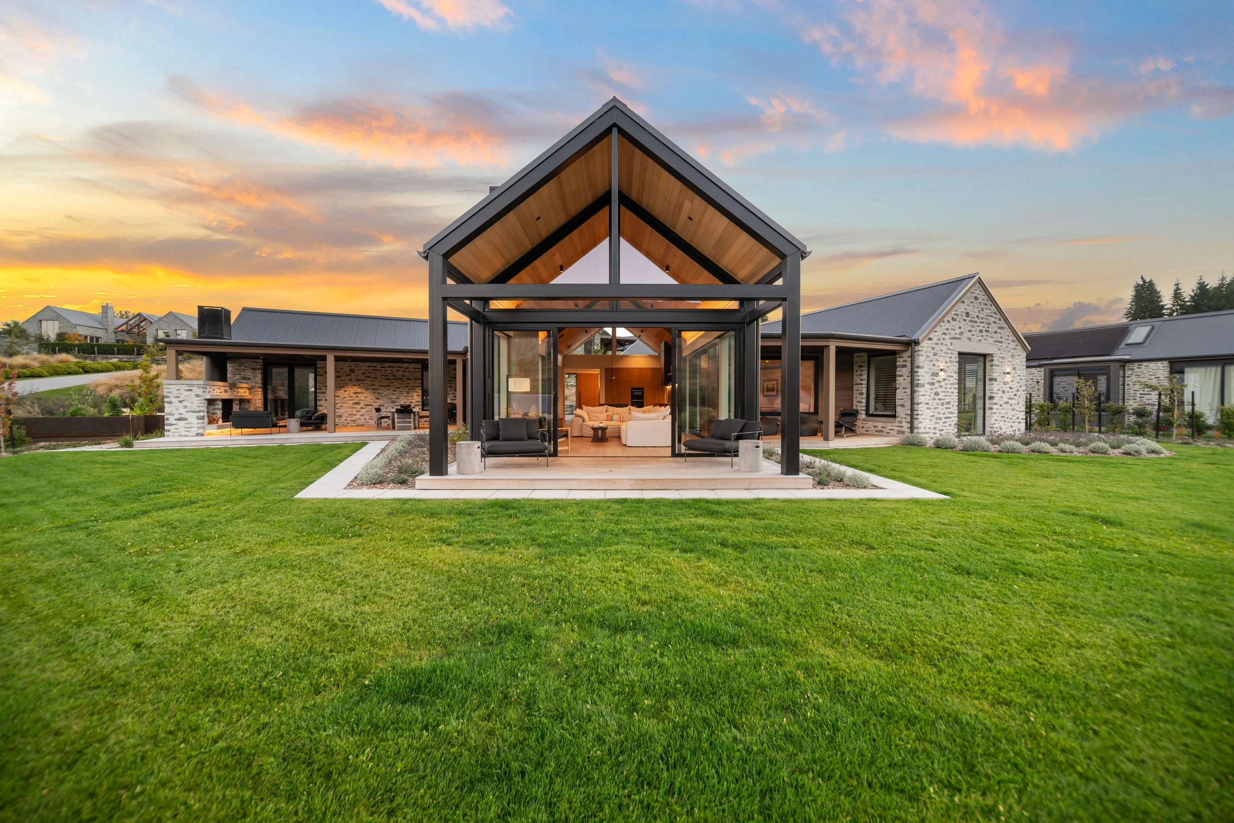 Modern house with glass walls and a metal roof, surrounded by a lush green lawn and garden, under a sunset sky with pink and orange clouds.