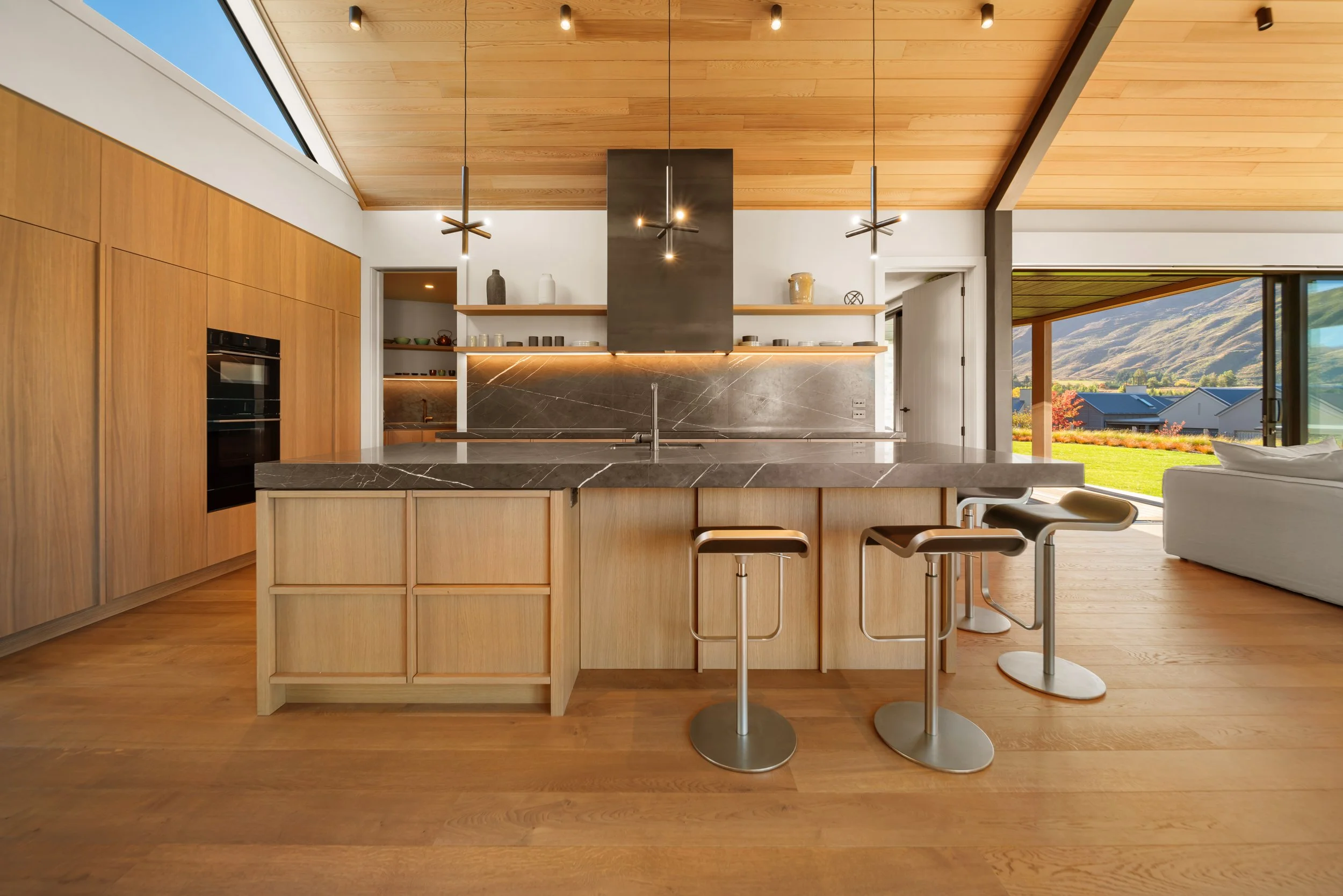 Modern kitchen with wood cabinetry, a black marble island, pendant lights, open shelves displaying dishes and decorative items, and large windows showing a view of mountains and houses.