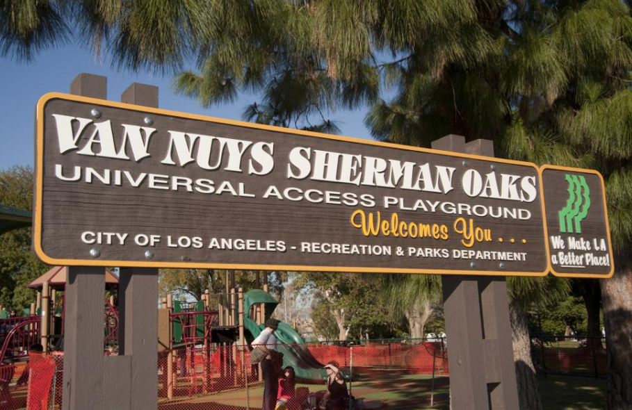 Sign for Van Nuys Sherman Oaks Universal Access Playground in Los Angeles, California, with playground equipment and children playing in the background.