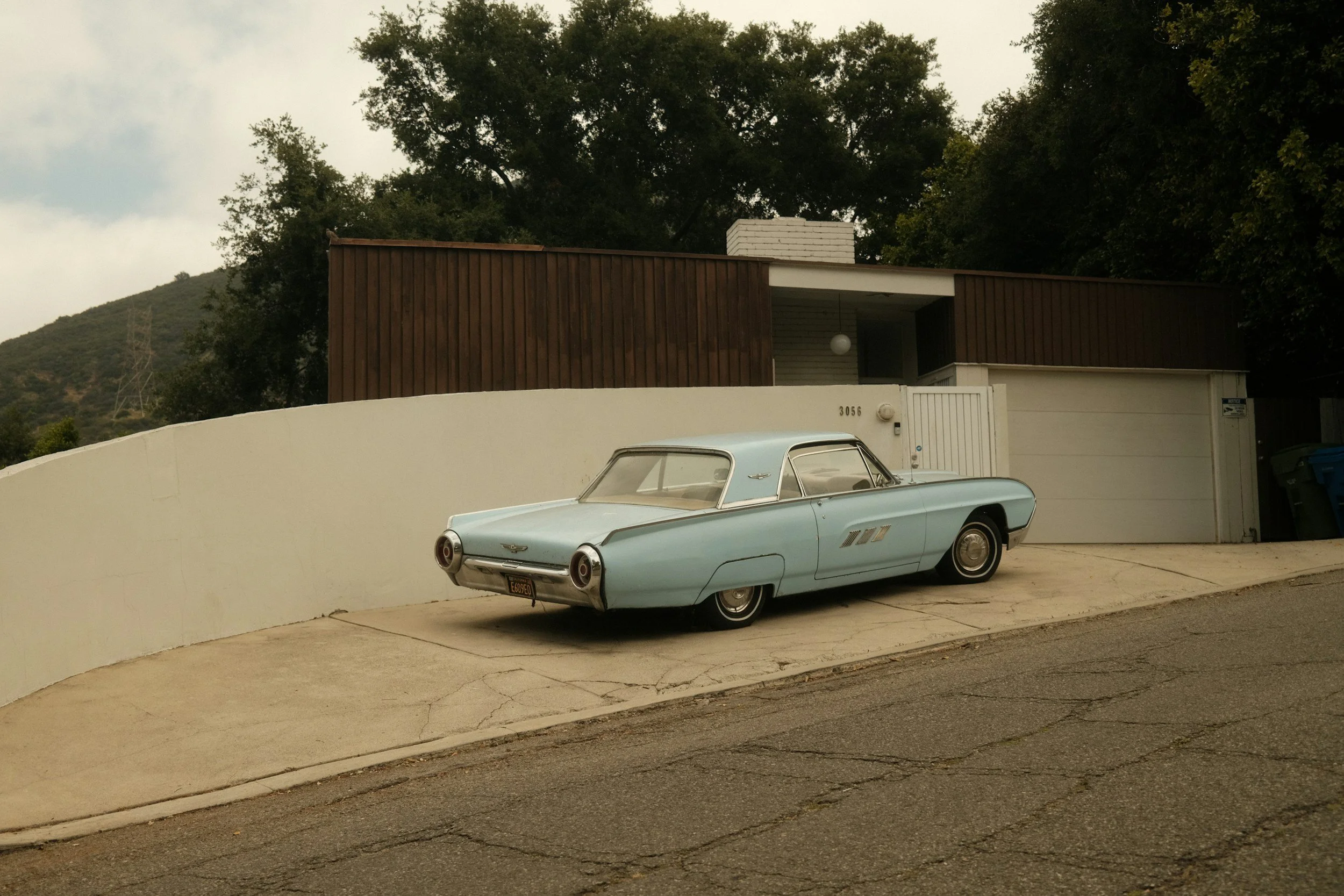 A vintage light blue car parked on a driveway in front of a modern house with wooden and white siding and a white wall.