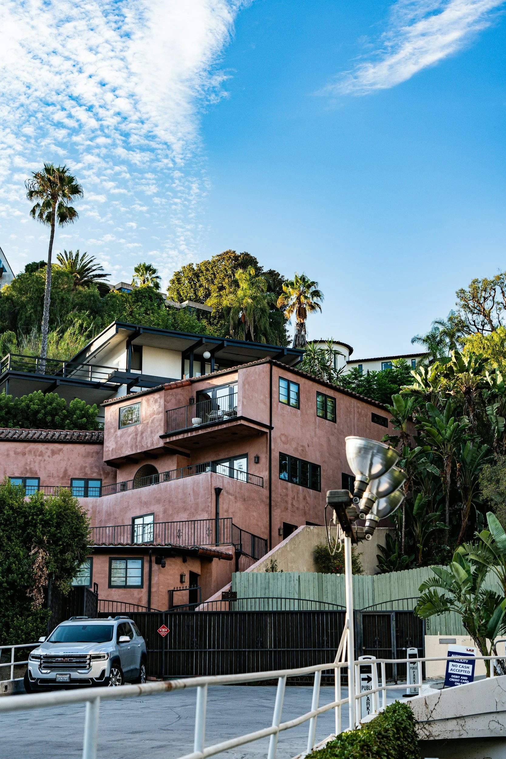 Residential hillside buildings with palm trees under a partly cloudy sky, parking area with car and parking payment machine.