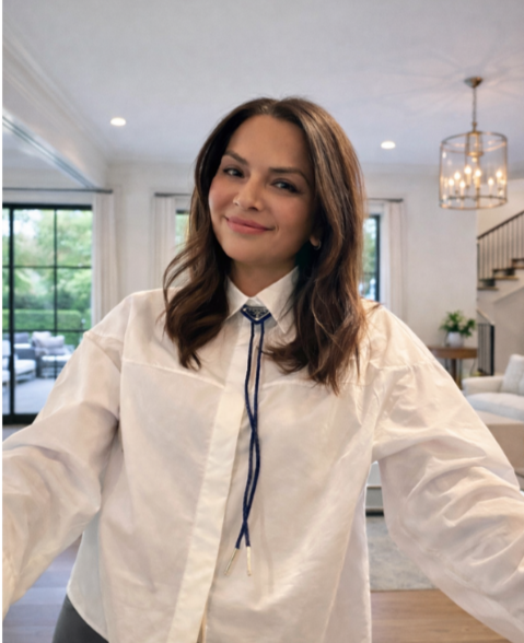 A young woman with brown hair smiling and taking a selfie in a bright, modern living room with large windows and a staircase in the background.