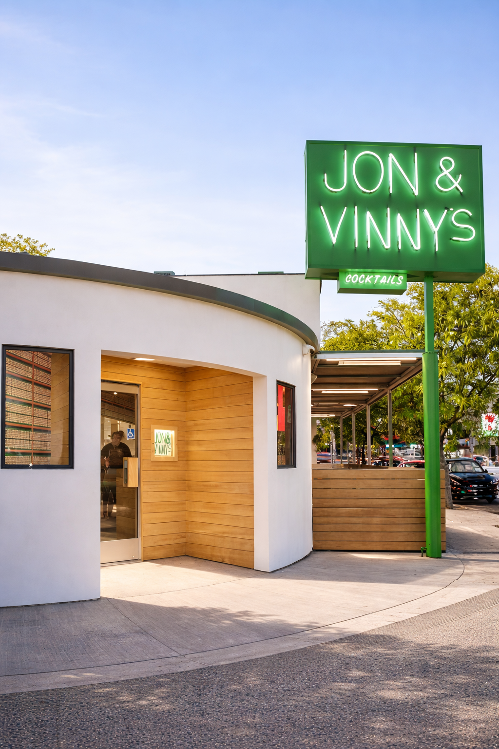 Exterior of Jon & Vinny's restaurant with a neon sign reading 'Jon & Vinny's' and a smaller sign below that says 'Cocktails', featuring a modern building design with white walls and wood accents, and trees in the background.