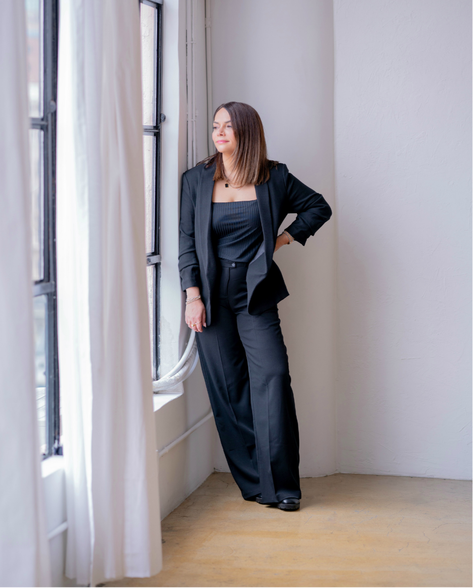 A woman in a black suit standing by a window with white curtains, looking outside with a contemplative expression.