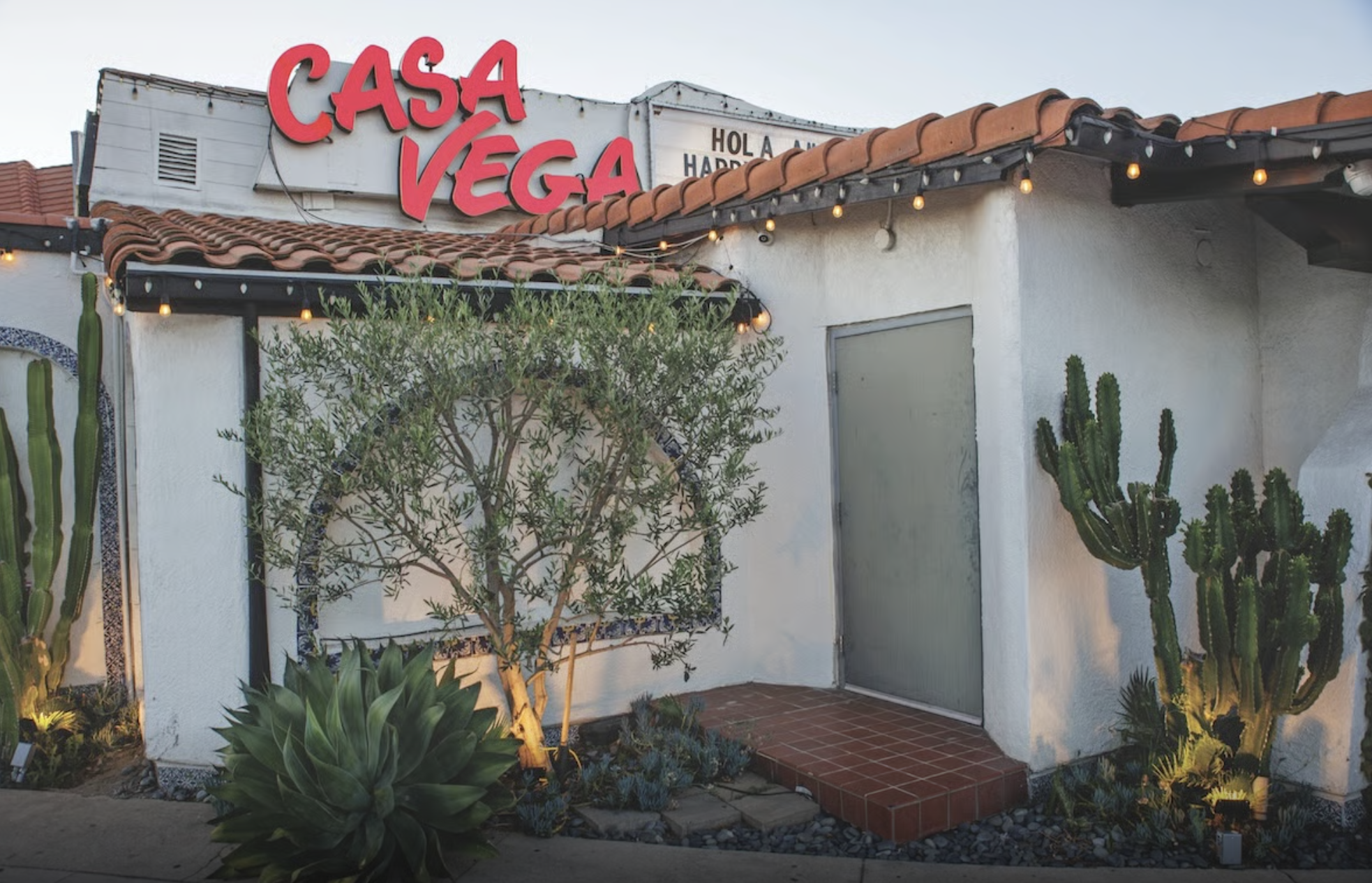 Exterior of a Spanish-style building with a white stucco wall, a red tile roof, and a sign that reads 'Casa Vega' in large red letters. There are lit string lights along the roof and desert plants, including cacti and succulents, in the foreground.