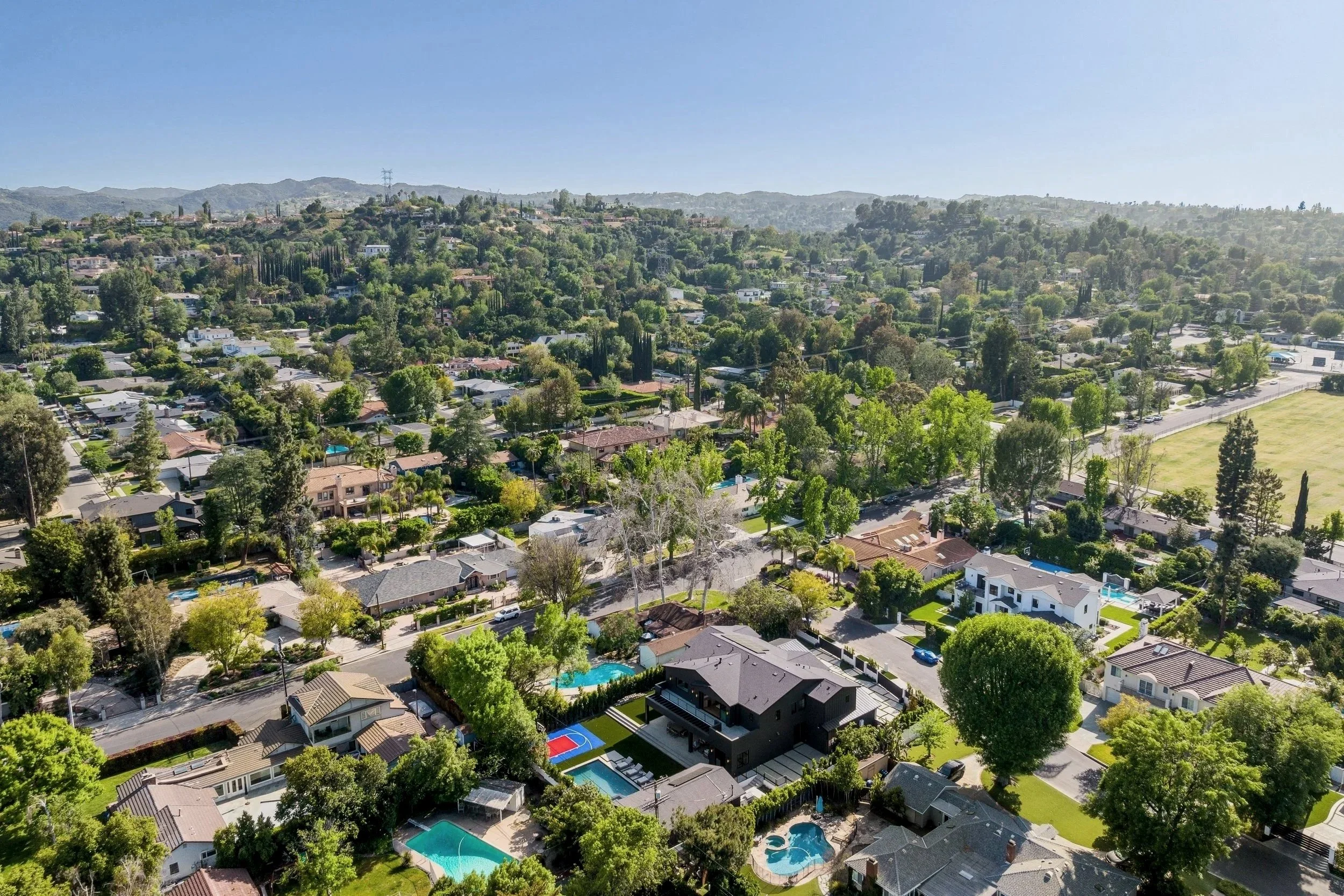 Aerial view of a suburban neighborhood with houses, trees, swimming pools, and mountains in the background under a clear blue sky.