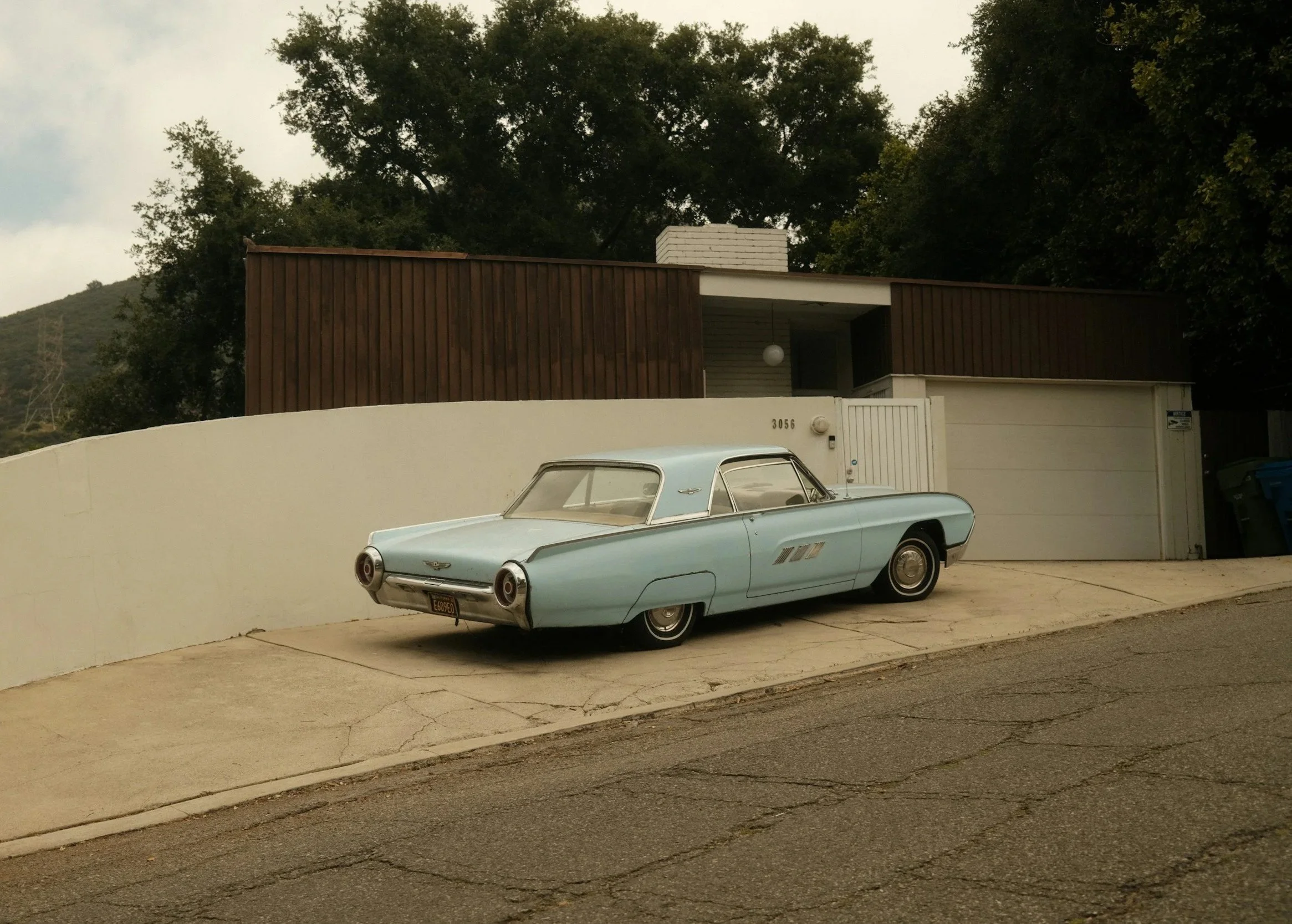 A vintage light blue car parked in front of a modern house with wooden siding, white walls, and a garage door. There are trees and hills in the background under an overcast sky.