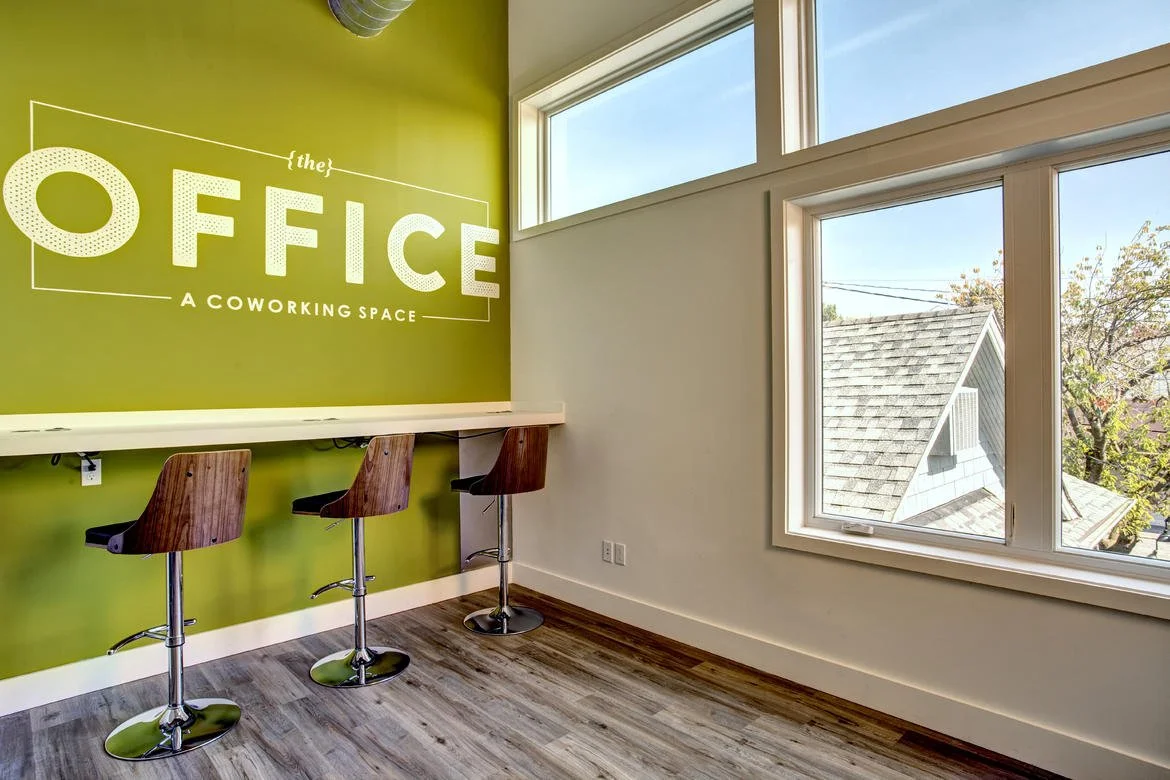 Interior of a coworking space office with a bright green wall, white wall with large windows, three wooden bar stools at a high counter, and sunlight coming through the windows.
