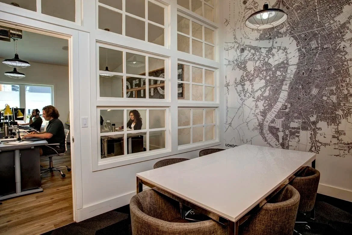 Office space with a meeting area showing a large white table and six chairs, a wall mural of a city map of London, and a glass partition separating it from a workspace with people working at desks.