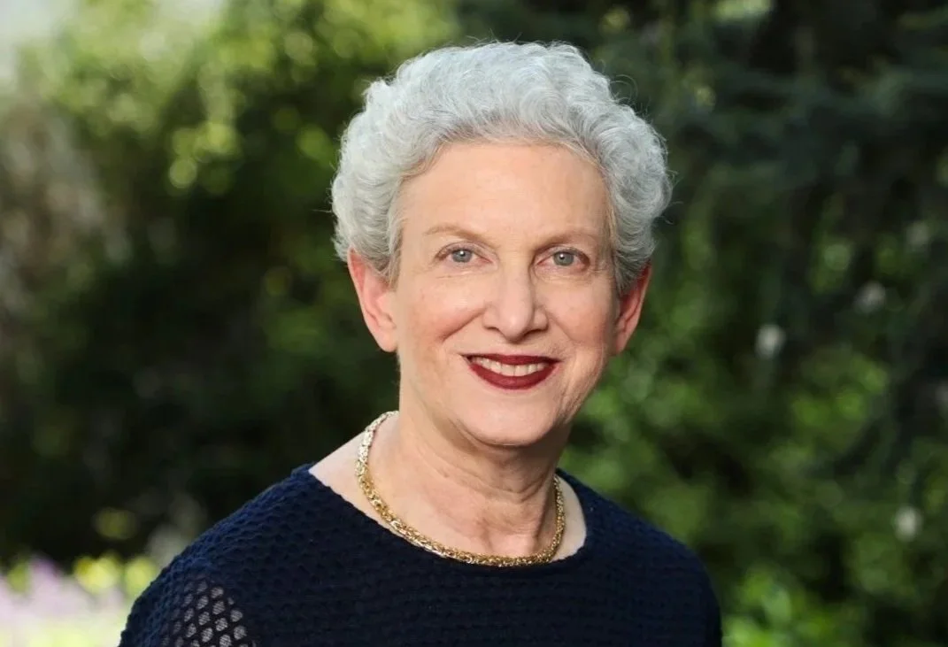 A portrait of an elderly woman with short white curly hair, wearing a dark marine sweater, a gold necklace, and dark lipstick, standing outdoors with green blurred foliage in the background.