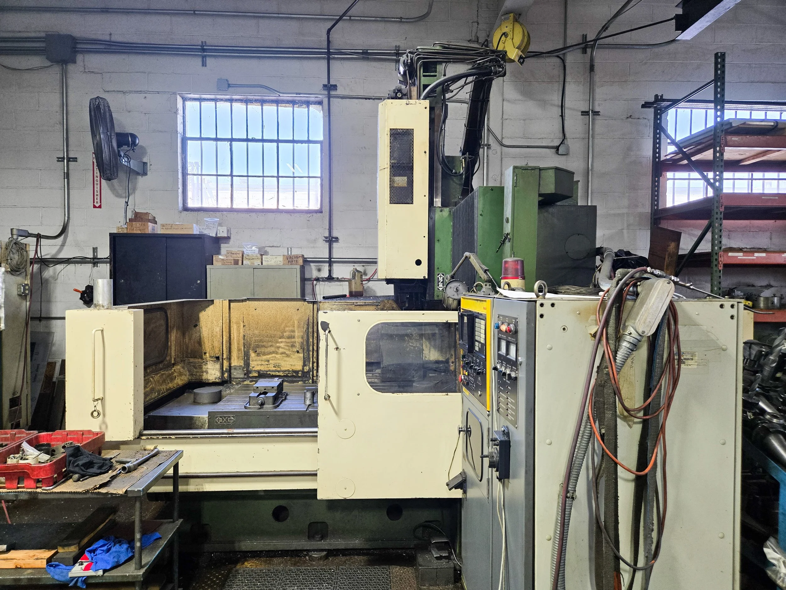 An industrial workshop with a large CNC machine in the center, surrounded by tools, boxes, and industrial shelving. Two windows provide natural light, and various wires and equipment are visible around the workspace.