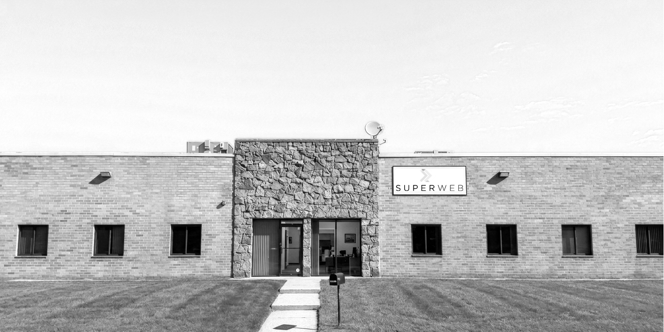 Front view of a single-story commercial building with a brick exterior, a stone façade around the entrance, a sign that reads "Superweb," four small windows, a sidewalk, and a grassy lawn.