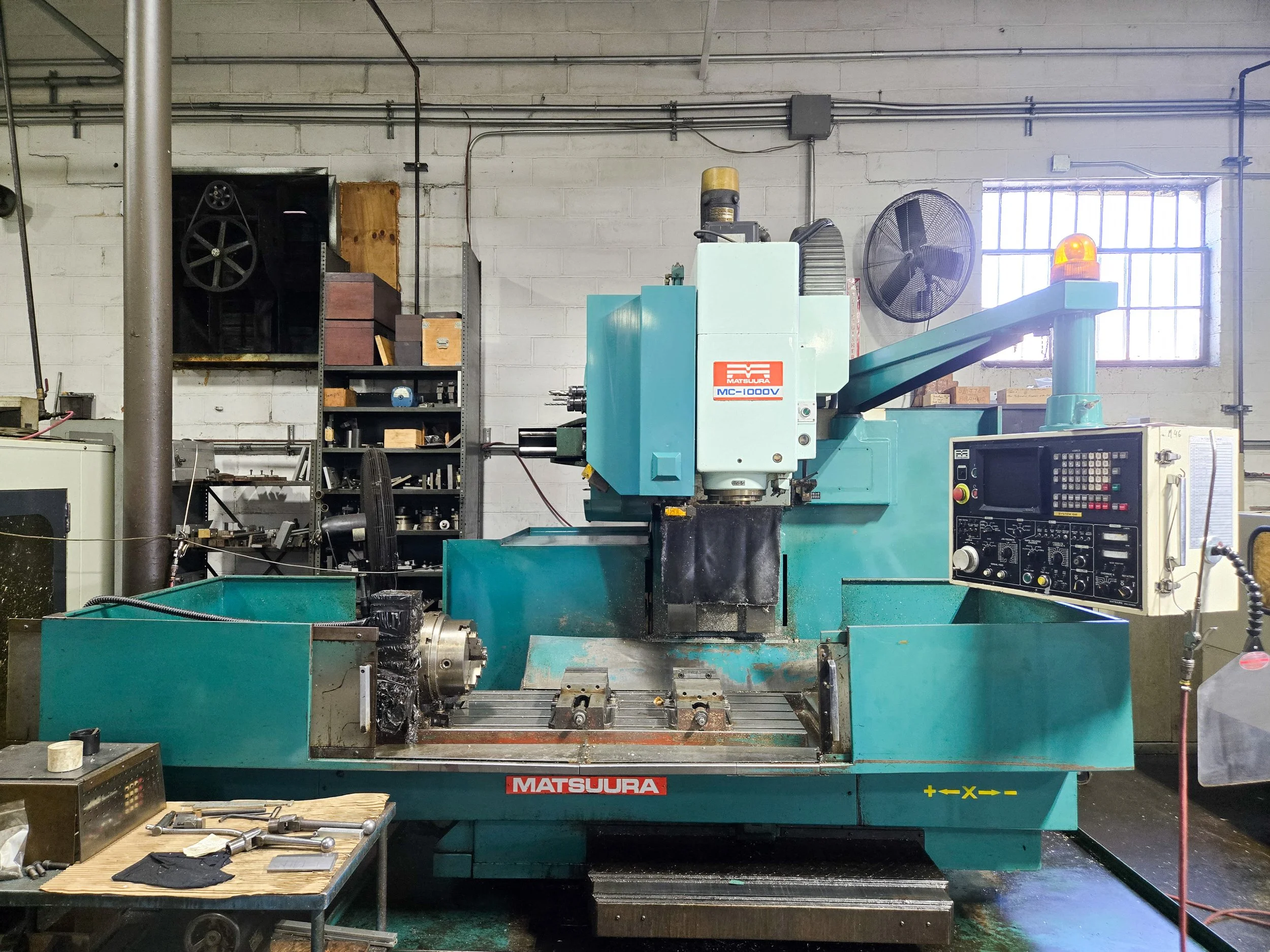 Industrial workshop with a teal-colored machine labeled 'Matsuura' in front of a wall with pipes and a window. There is a worktable with tools and materials, a black fan, and an orange warning light on the machine.