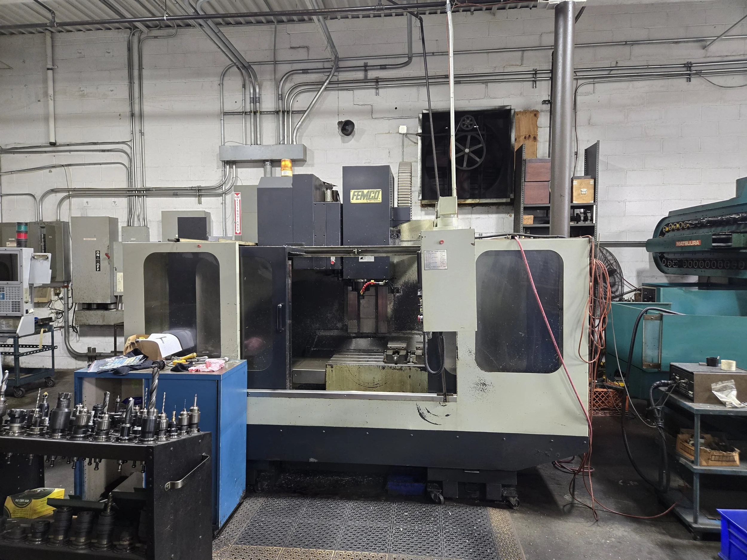 Industrial workshop with machinery, metal pipes on walls, a rack with stapled boxes, and metal parts on tables.