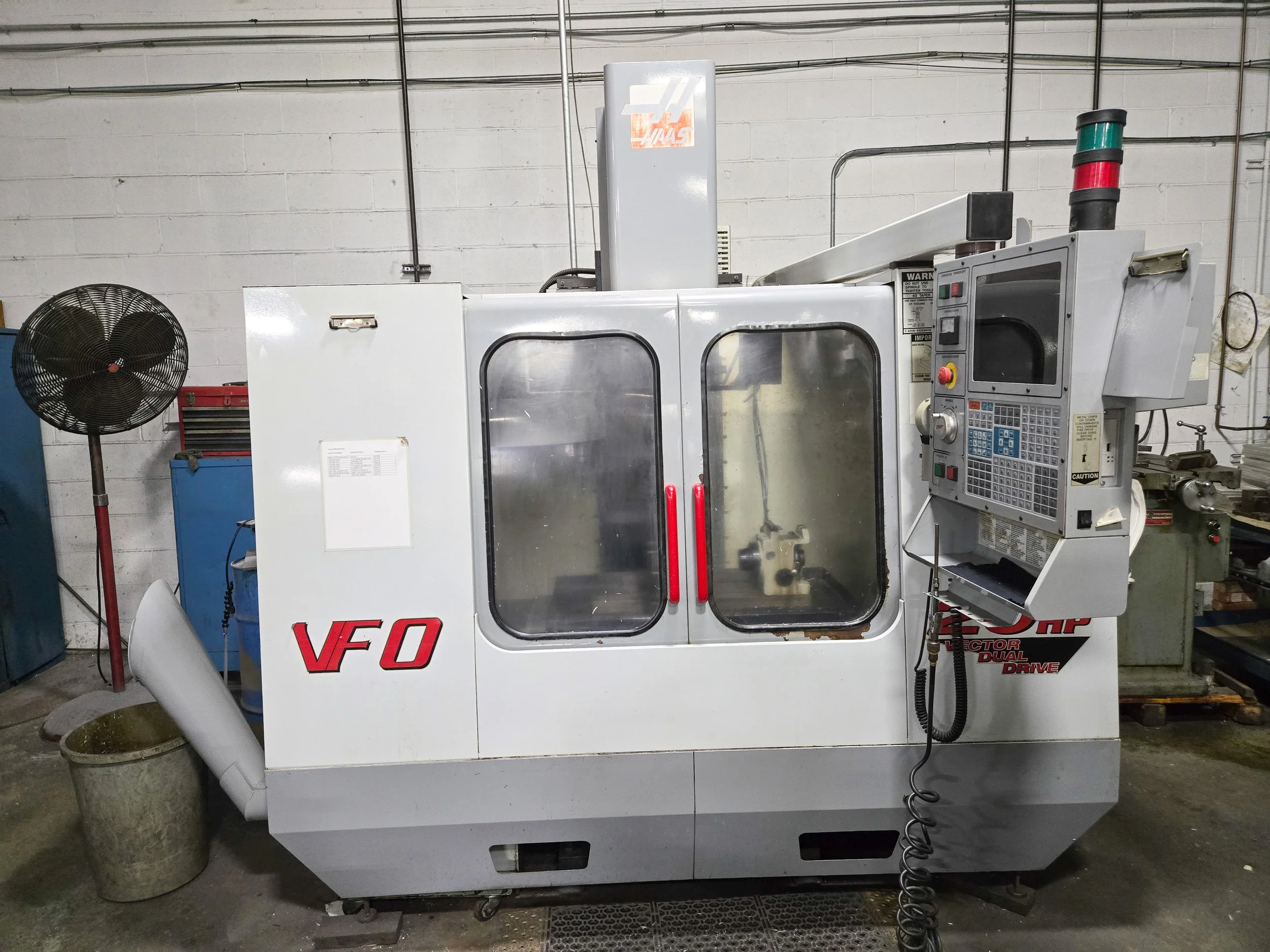 Industrial machinery in a workshop, featuring a large white metal machine with a control panel, warning labels, and red and green indicator lights. A black fan stands to the left, and various tools and containers are in the background against a cinde
