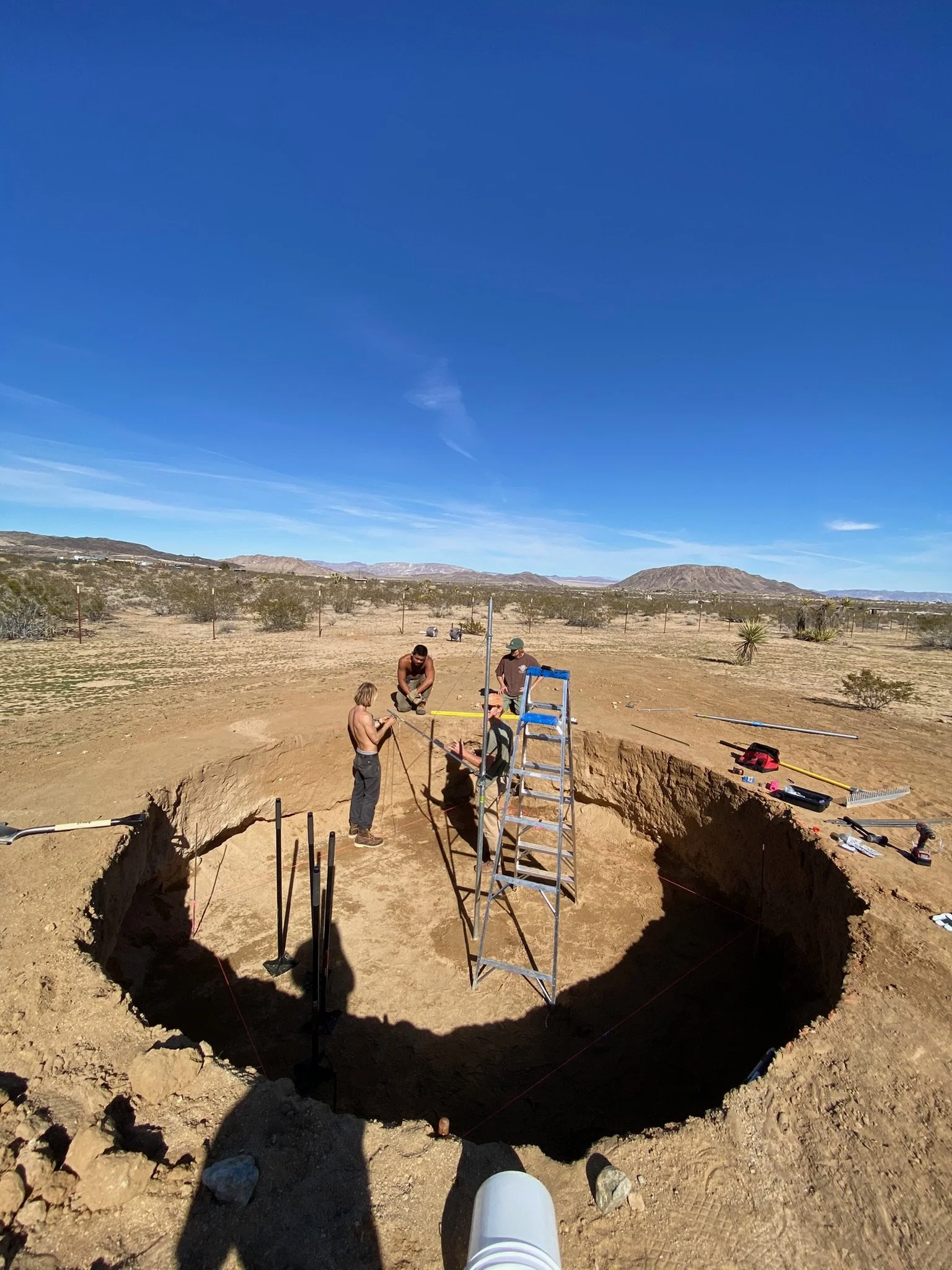 People excavating and working inside a large hole in the desert with tools, ladder, and equipment around.