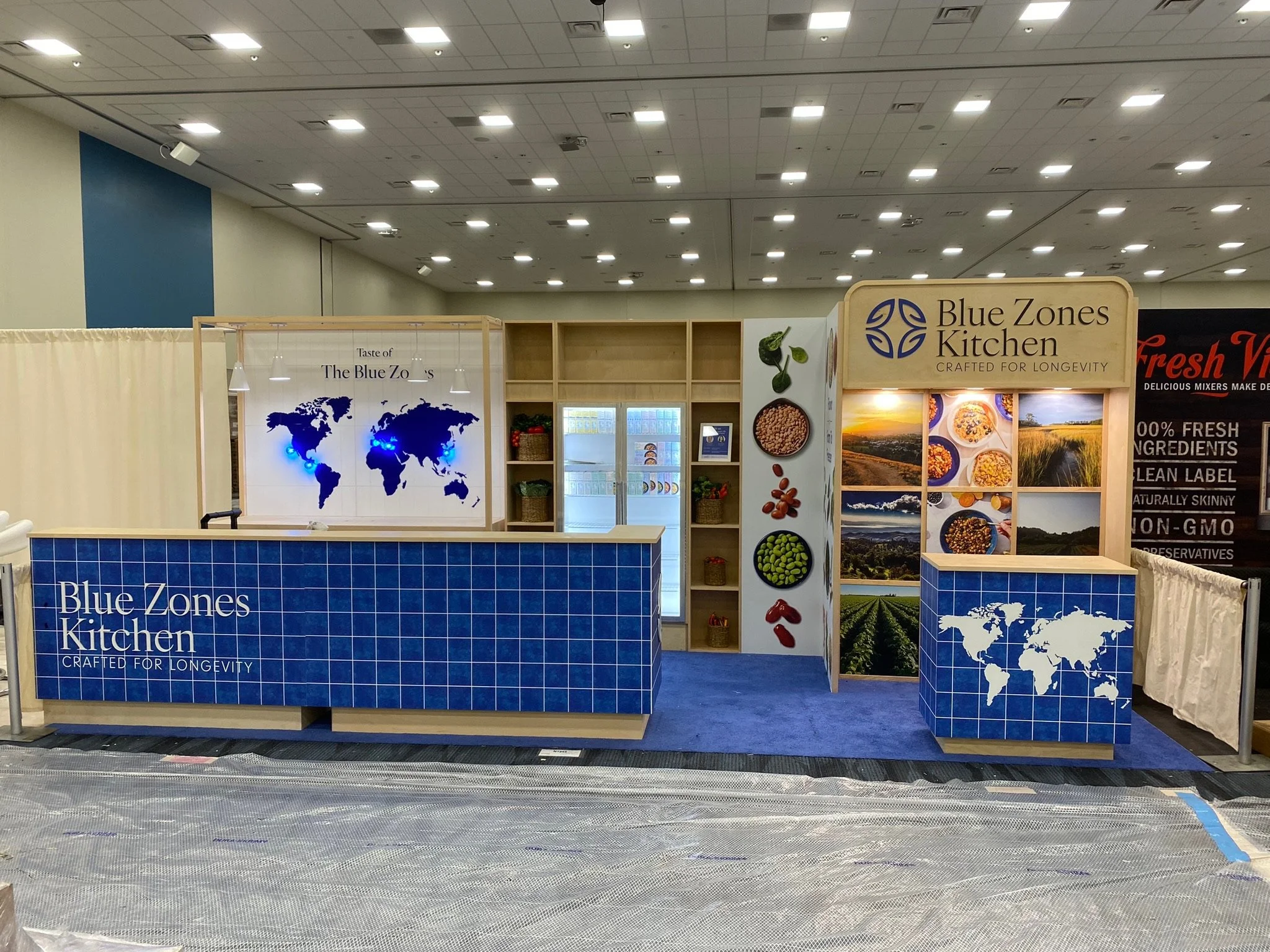 Exhibition booth for Blue Zones Kitchen featuring a blue tiled counter, world map graphics, and images of healthy foods and landscapes.