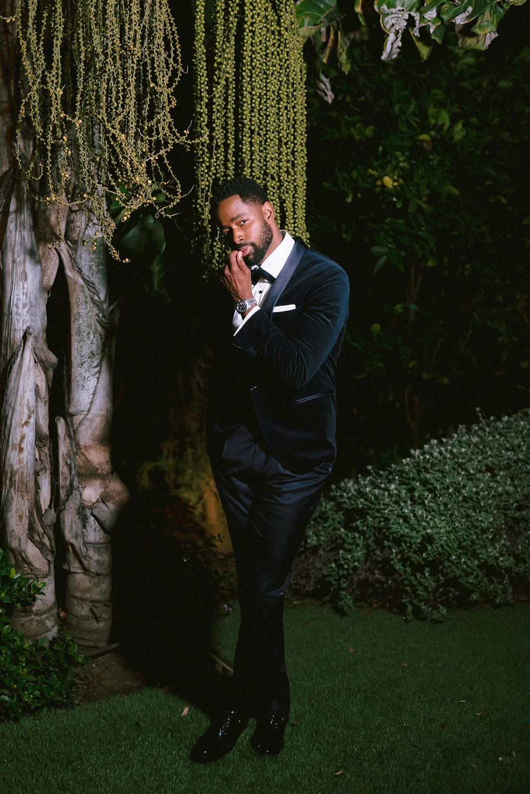 A man in a black tuxedo with a white shirt and bow tie, standing outdoors at night, posing with his hand near his face amidst greenery and hanging decorative plants.