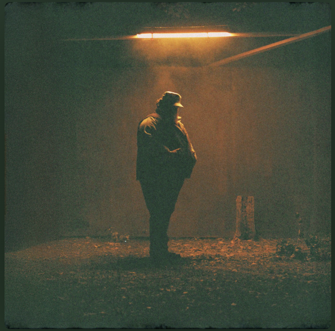 A person standing alone in a dimly lit, foggy outdoor setting under an orange streetlamp at night.