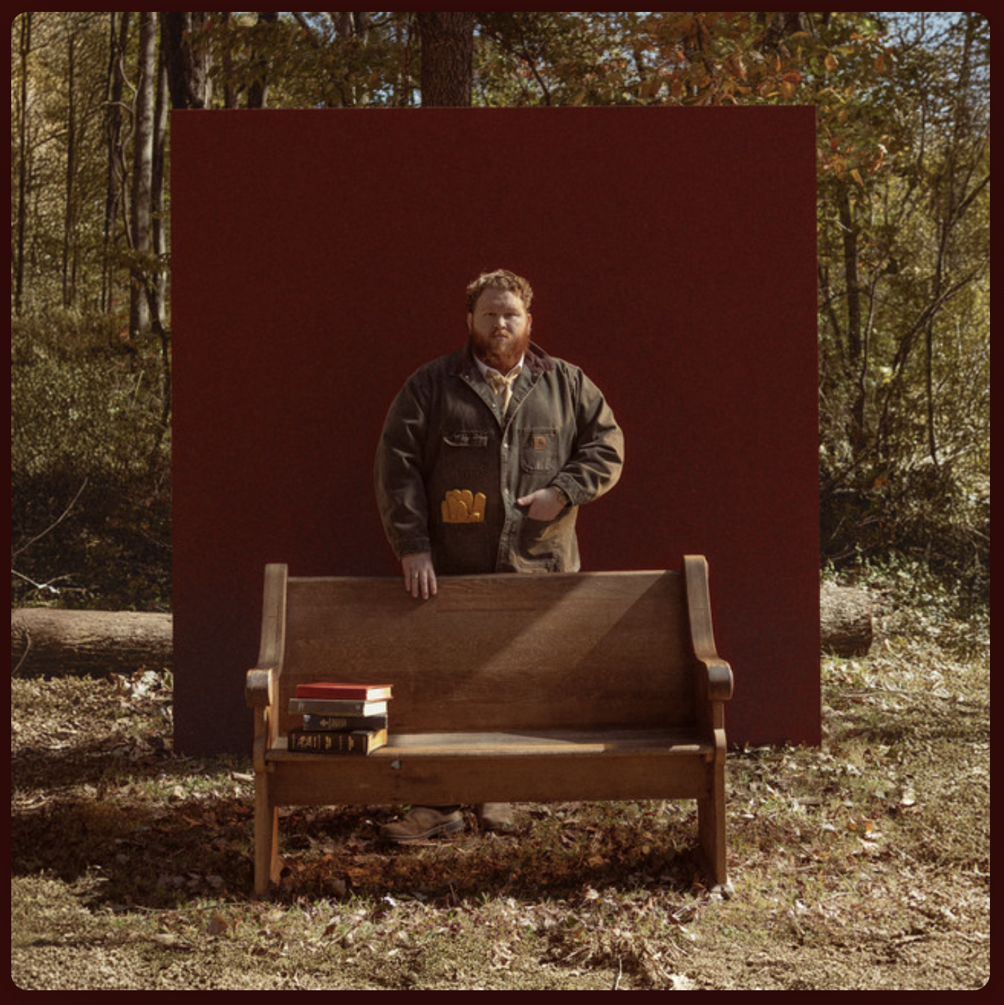 A man with a beard standing outdoors behind a wooden bench with books on it, in front of a red backdrop, in a forested area with autumn leaves.