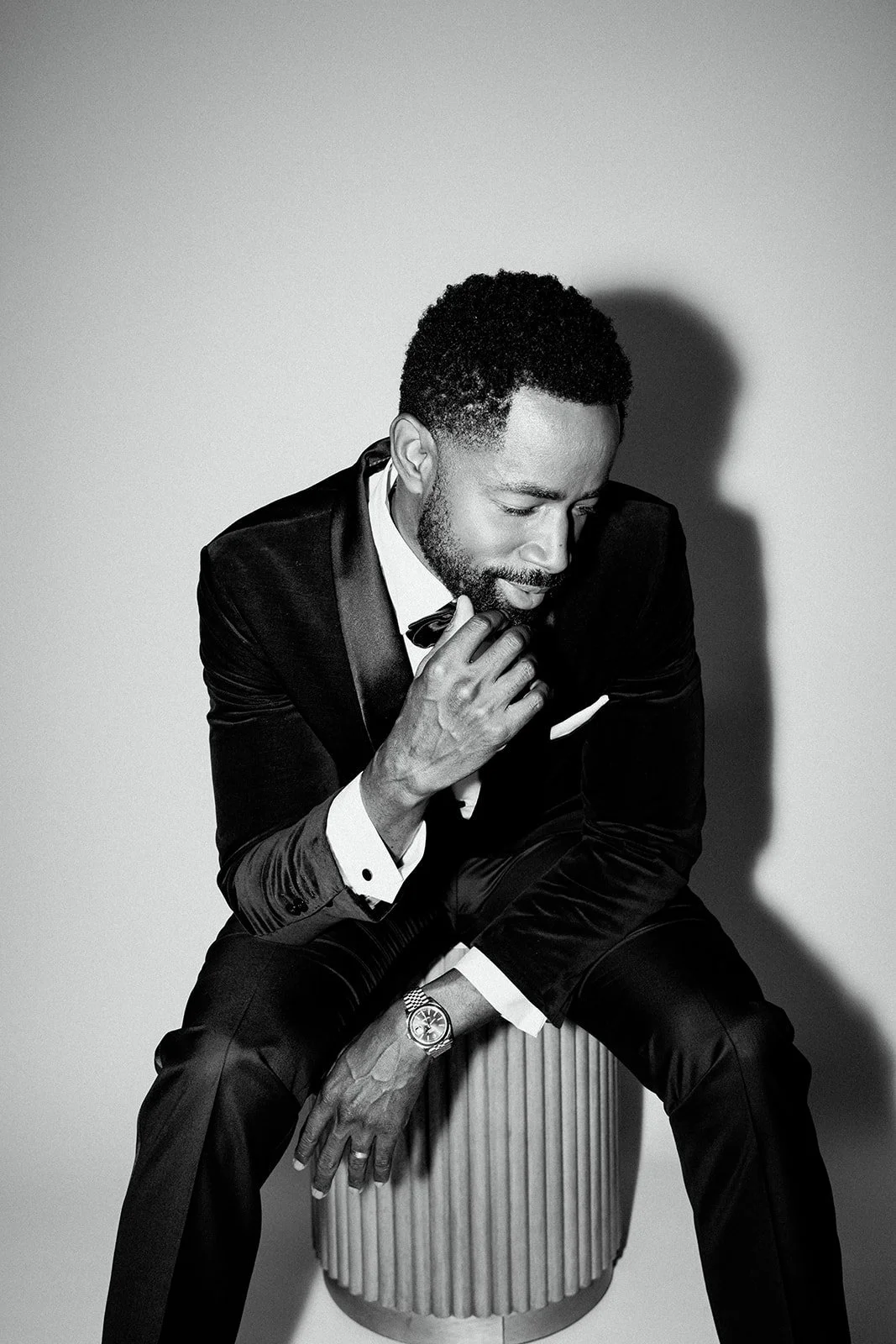 A man in a tuxedo sitting on a cylindrical stool, looking down while touching his chin with his right hand. Black and white photograph.