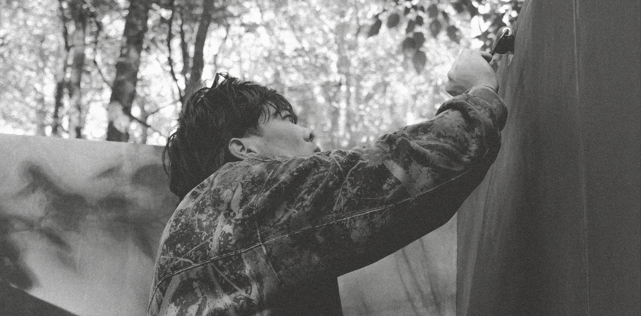 A person with short, dark hair and wearing a camouflage jacket is climbing a wall with the help of climbing holds in an outdoor forest setting.