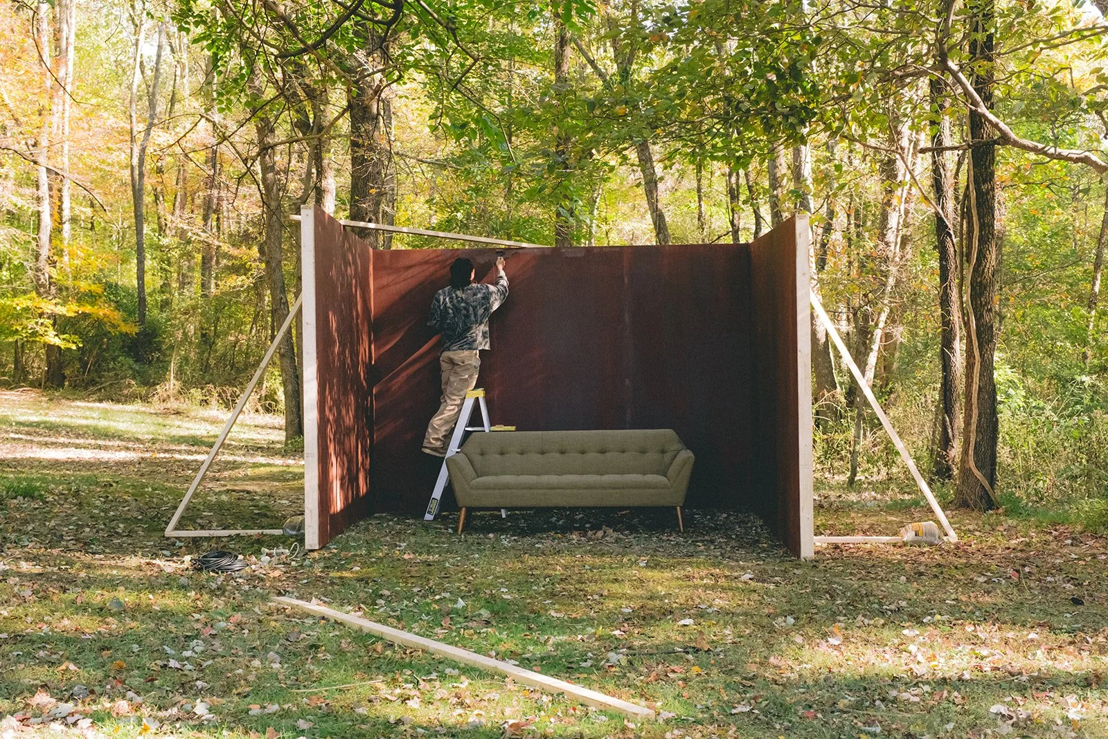 Man standing on a ladder adjusting a large brown partition wall outdoors in a wooded area. A green couch is placed against the wall, and trees and autumn foliage are visible in the background.