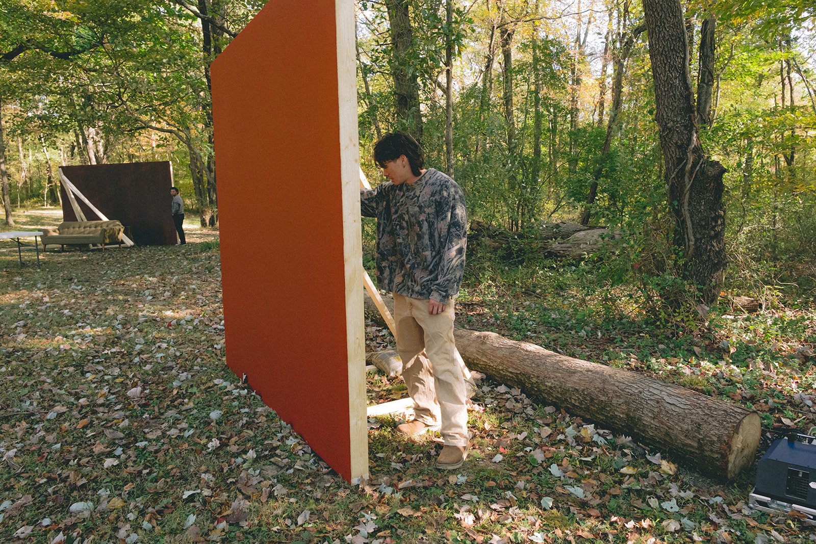 A young man with dark hair, wearing a camo-patterned shirt and beige pants, stands outdoors in a wooded area during daytime. He is touching a large, orange, vertical panel that is set up in the middle of the scene. There is fallen leaf-covered ground, a log on the right, and a series of furniture, including a couch and a partition, towards the background.