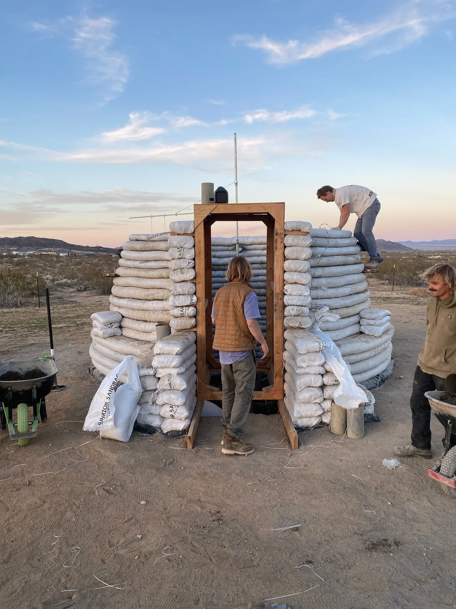 Construction of a small structure using sandbags in a desert landscape at sunset, with three men working around it.