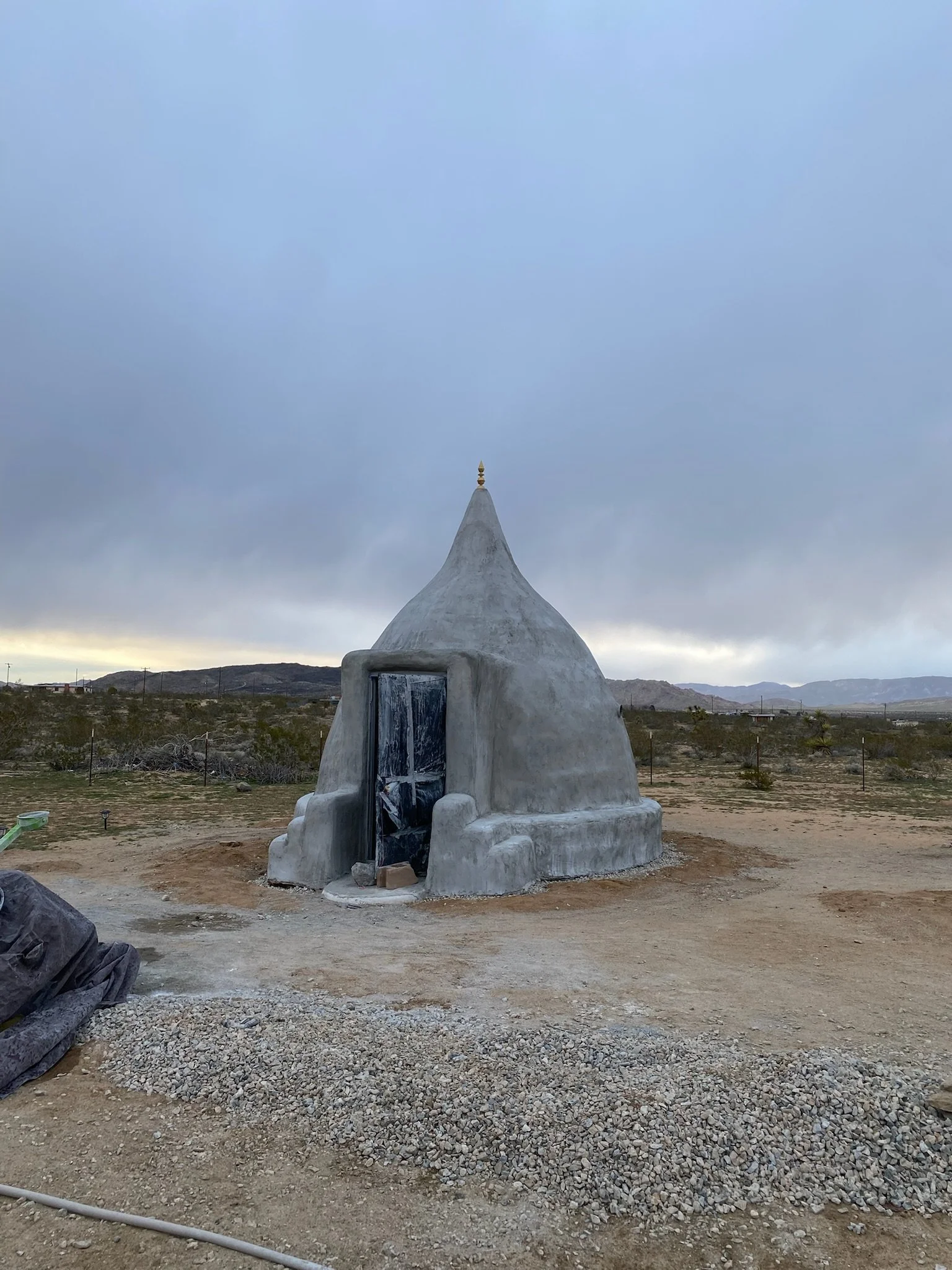 Small, dome-shaped structure resembling a mud hut with a pointed top, located in an arid, flat landscape with mountains in the distance and cloudy sky overhead.