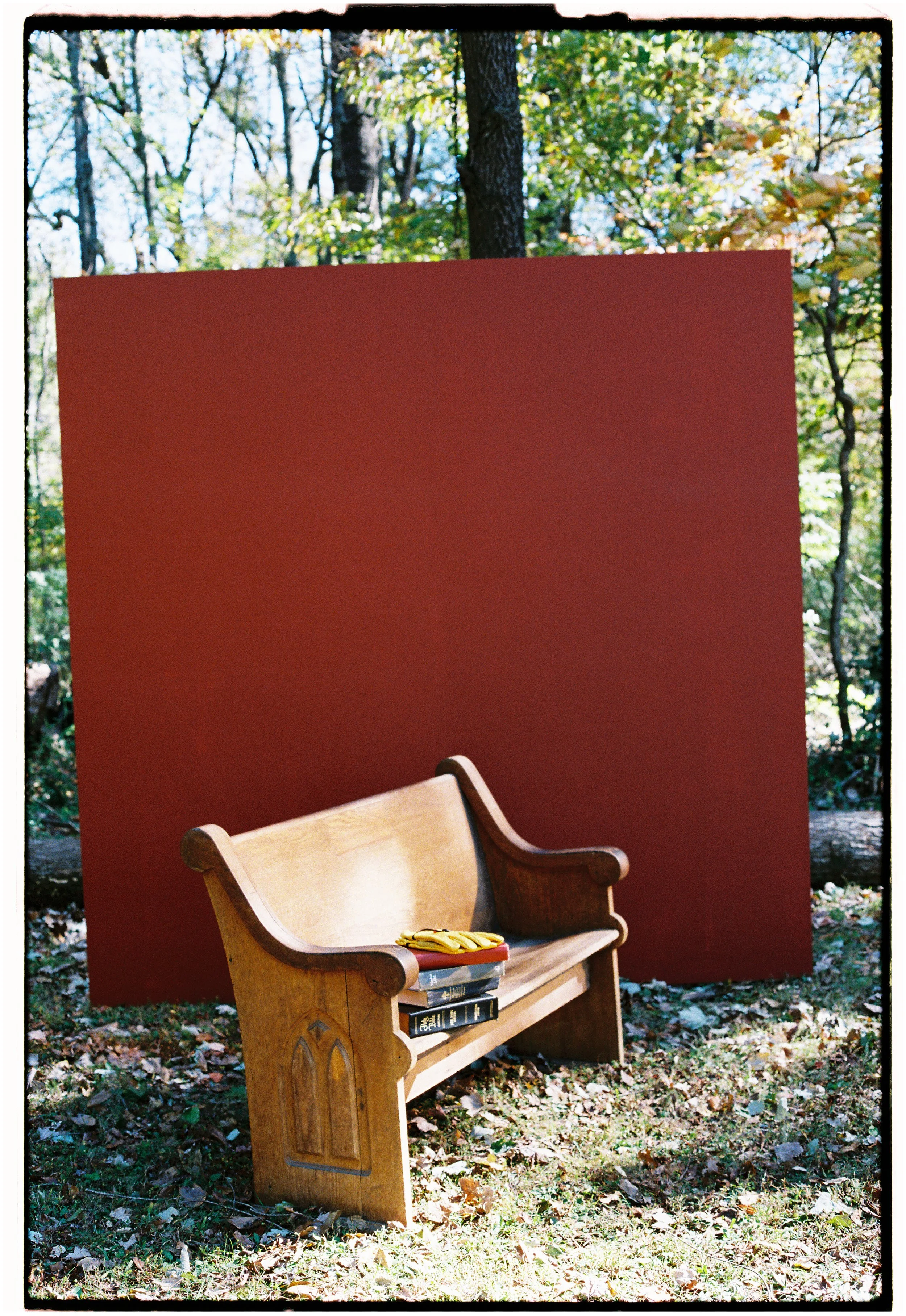 A wooden church pew with books and yellow gloves on the seat, set outdoors on grass with fallen leaves and a red backdrop, trees and blue sky in the background.