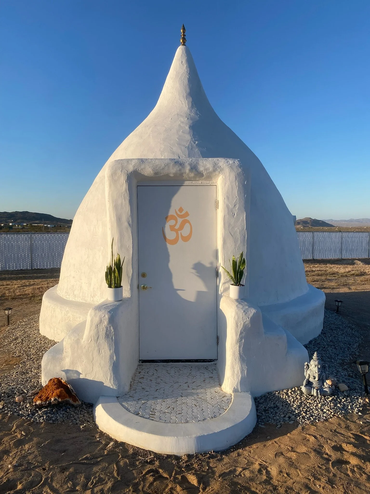 Small white, cone-shaped building with a pointed roof, decorated with an Om symbol on the door, flanked by potted plants, and a small statue of Ganesha nearby, set in an outdoor sandy area with a fence and hills in the background.