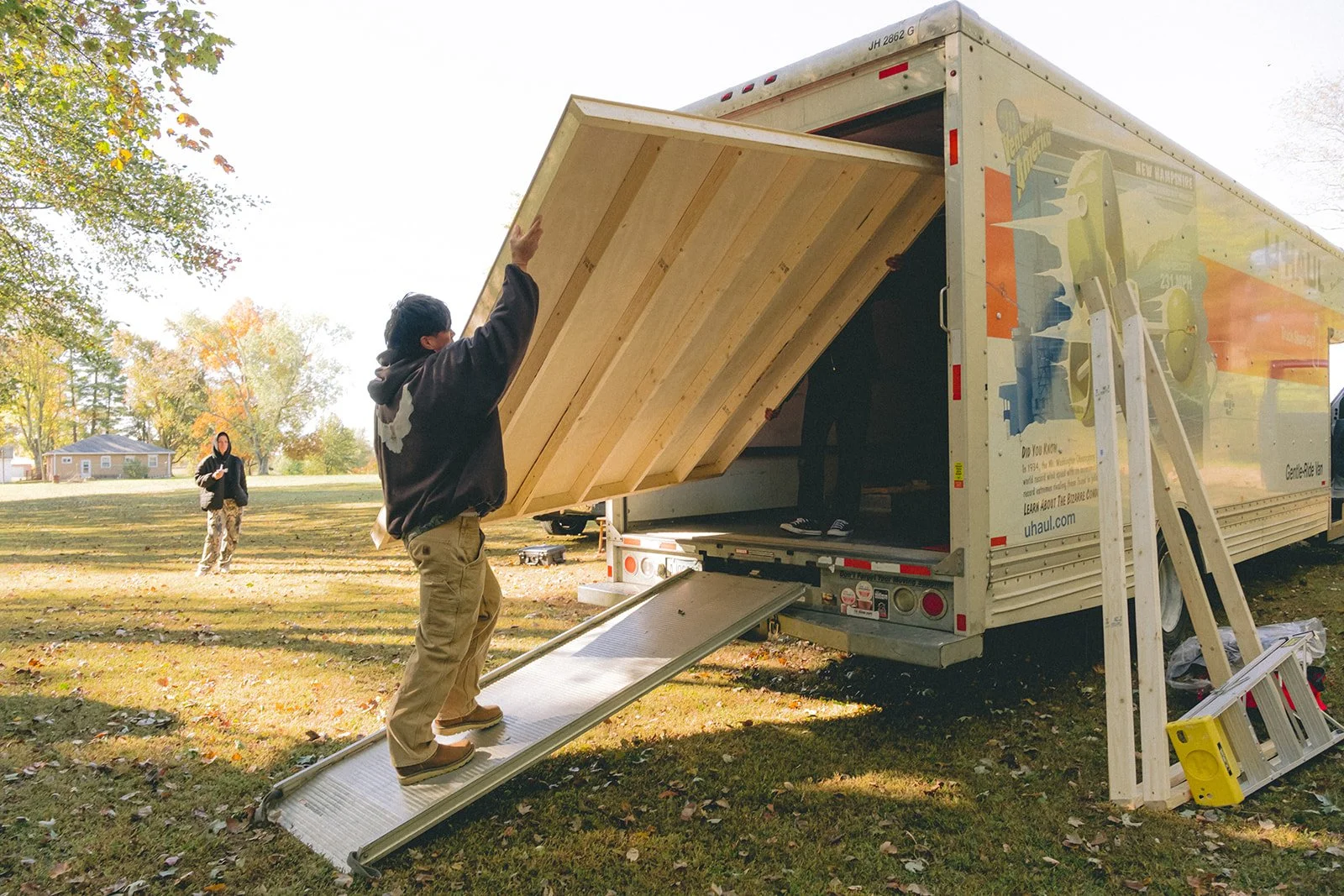 Two people are loading a large wooden piece into a U-Haul truck using a ramp, with autumn trees in the background.