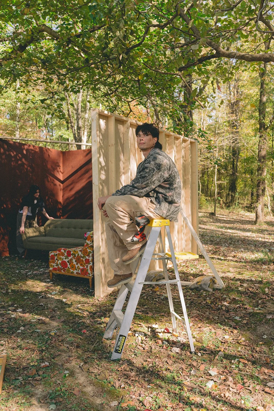 A man sitting on a step ladder in a forest, with partial construction of a wooden wall behind him. There is a woman inside the structure, arranging furniture, and a floral sofa and a green sofa are visible inside. Trees and foliage surround the scene.